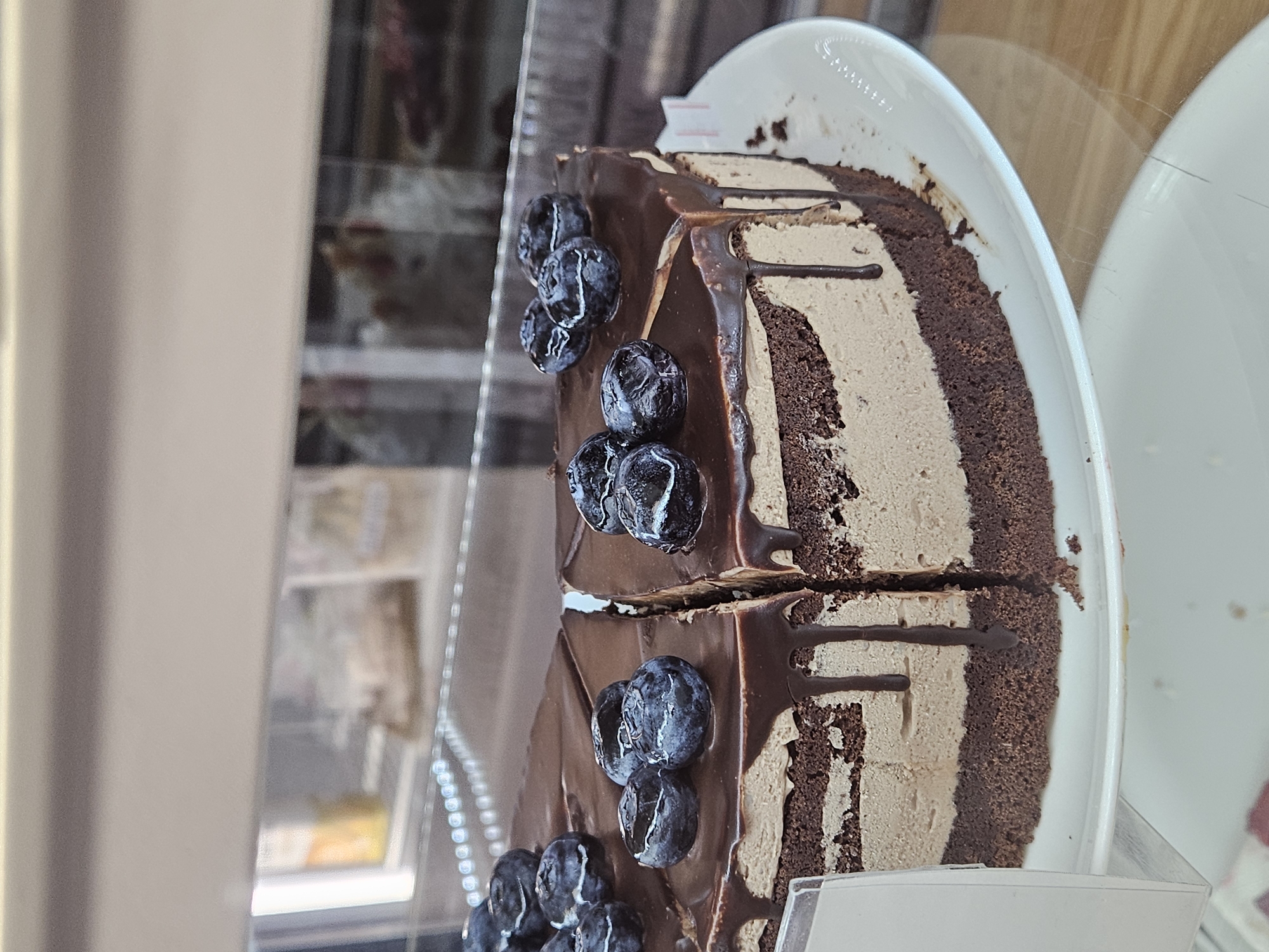 A close-up, rotated shot captures several slices of a decadent chocolate cake displayed in what appears to be a refrigerated case at a bakery or cafe. The cake is composed of rich, dark chocolate cake layers alternating with lighter, creamy, possibly mocha or coffee-flavored, mousse or frosting layers.
The top and sides of the cake slices are generously coated in a glossy, dark chocolate ganache or glaze, which drips attractively down the sides. Scattered on top of the chocolate glaze are fresh, plump blueberries, some appearing slightly wrinkled, adding a touch of color and natural sweetness.
The cake slices are presented upright on pristine white ceramic plates. At least three slices are clearly visible, with the first two sharing one plate and a portion of a third slice visible on another plate further to the right. The cake is seen through a clear protective barrier, likely glass or plastic, which reflects some ambient light and shows slight smudges or scratches.
In the blurred background, through the clear barrier, faint outlines of other items or shelves can be discerned, consistent with a retail food display. A small, white rectangular label holder is partially visible at the bottom right, but no text on it is legible. The overall impression is one of an enticing dessert ready for consumption.