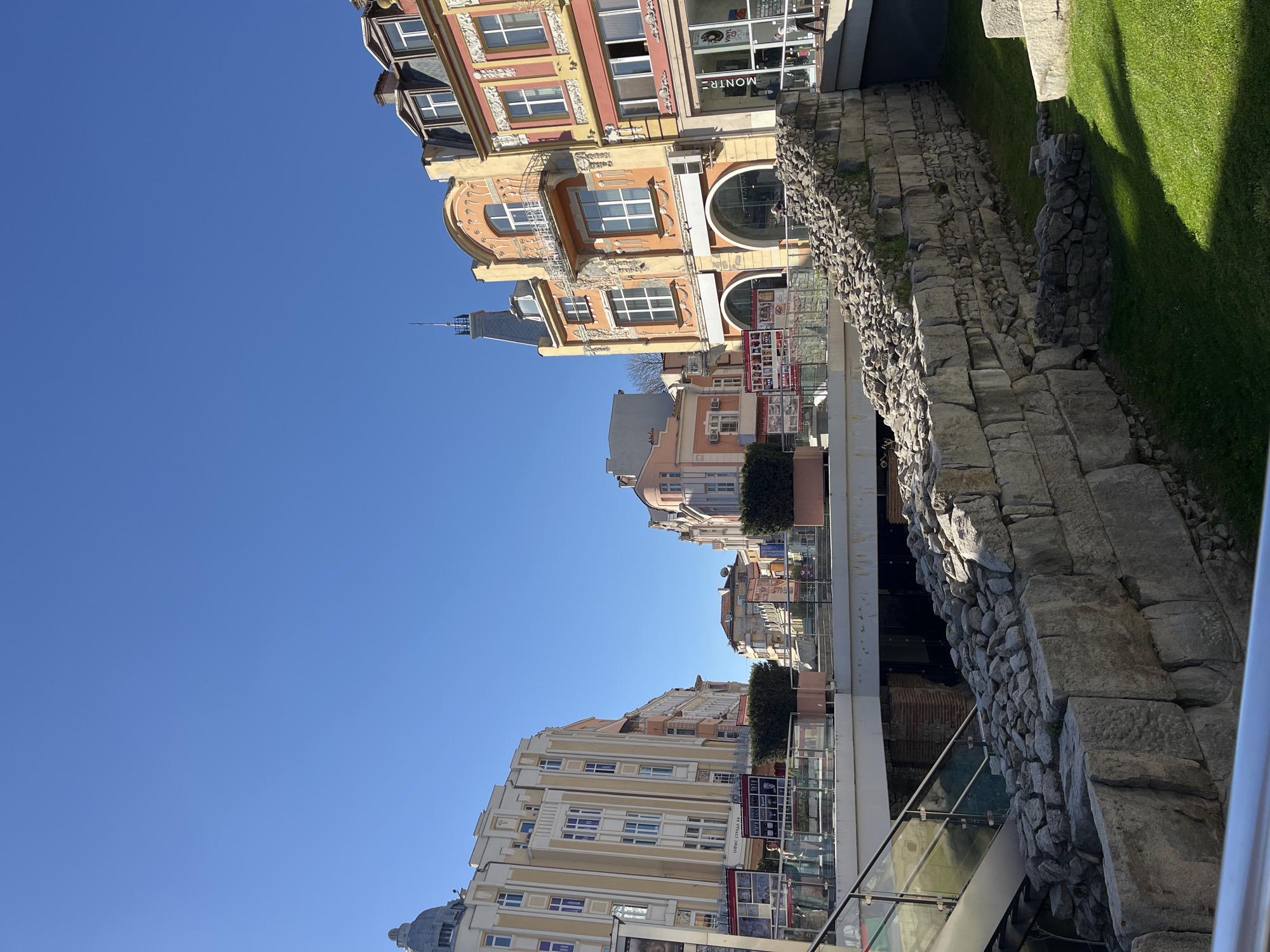 The image shows a street scene in Plovdiv, Bulgaria, featuring well-preserved Roman ruins adjacent to modern buildings. A stone wall, likely part of the ancient Roman city, runs along the right side, next to a grassy area. Adjacent to the wall is a modern glass-paneled walkway or structure, contrasting with the antiquity of the ruins. The buildings on the left side are a mix of architectural styles, with some appearing older and others more contemporary, suggesting an integration of historical and modern elements in the city. The sky is clear and blue. There are no people visible in the frame.