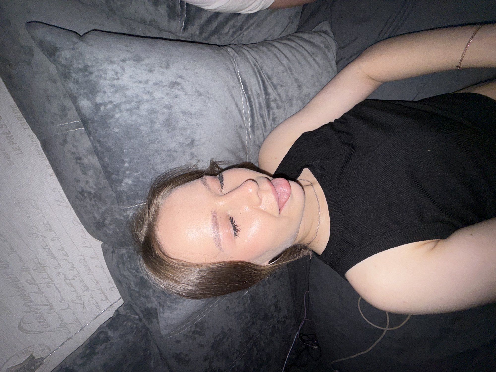 The media file shows a young woman lying on a grey sofa. She is wearing a black, ribbed tank top and a delicate silver bracelet on her right wrist. Her eyes are closed, and her tongue is sticking out slightly, suggesting a playful or relaxed mood. Her hair is brown and falls around her face and shoulders. The sofa has a textured, plush surface, and a patterned wallpaper with script is visible in the background, to the left of the sofa. There are also some wires or cables visible on the sofa near the woman. The lighting suggests it might be nighttime or indoors with artificial light. The overall scene appears to be a casual, personal moment within a home setting. No specific event or activity is clearly depicted, other than the woman resting or posing for the photo.