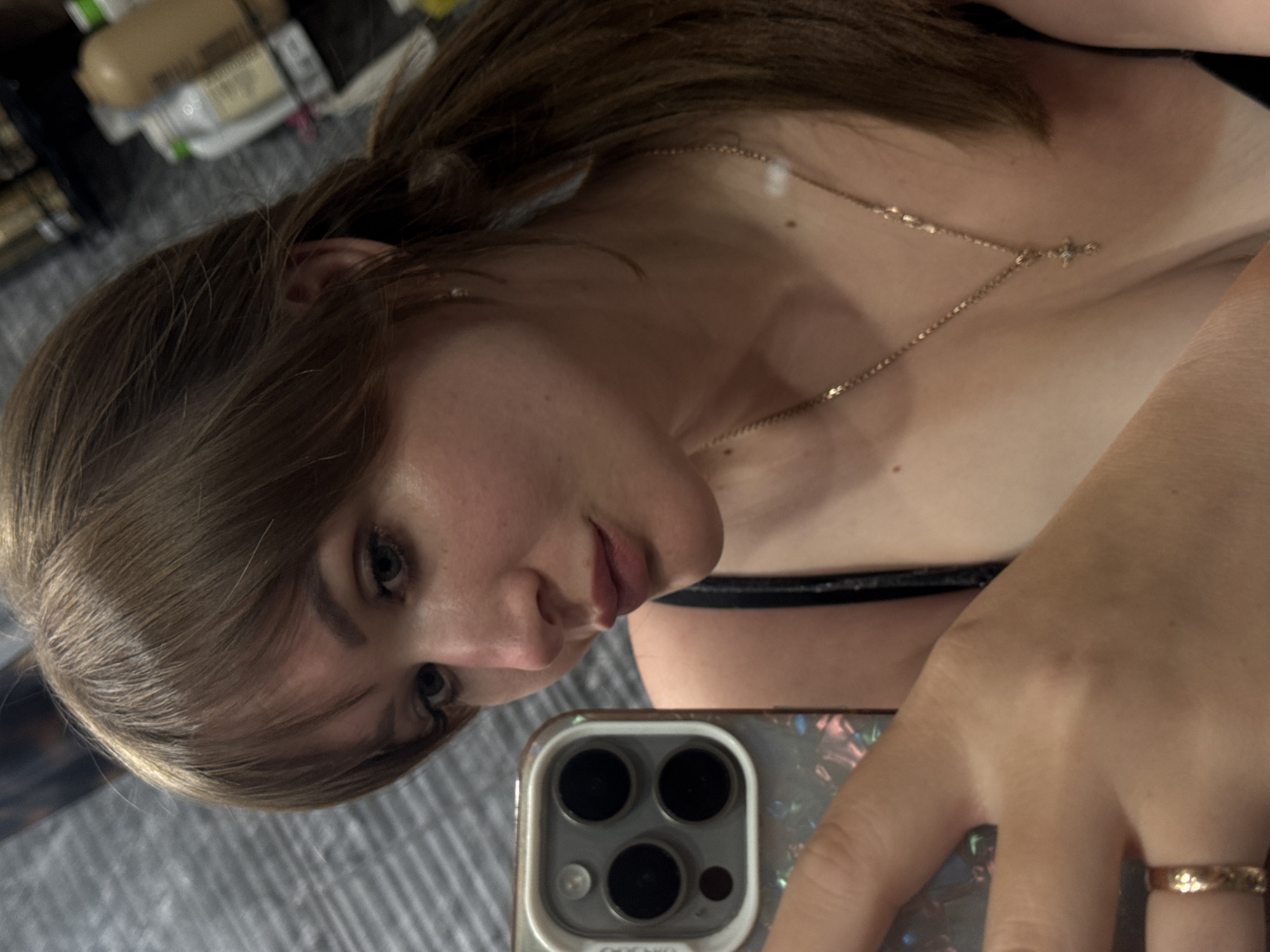 An angled selfie captures a young woman looking directly into the camera, with a slight, neutral expression. Her head is tilted, and her gaze is directed towards the lens. She has medium brown hair with bangs framing her face, and light-colored eyes enhanced with eyeliner. Her lips are slightly parted. She is wearing a dark top or bra and a delicate, gold-toned chain necklace with a small cross pendant. A simple gold-toned ring with an intricate pattern is visible on her right ring finger, which is holding a smartphone.
The smartphone appears to be an iPhone, identifiable by its triple camera module on the back, and it's protected by an iridescent or holographic case. A faint reflection of her face can be seen on the phone's screen. The background is blurred, showing shelves lined with various bottles, suggesting an indoor, personal setting like a bedroom or bathroom. The surface beneath her is a grey, textured material, possibly a carpet or fabric. The image is taken in a casual, self-portrait style. Faint text, possibly "oocoo," is discernible on the phone case. This scene is set in Chaikovsky, Russia.