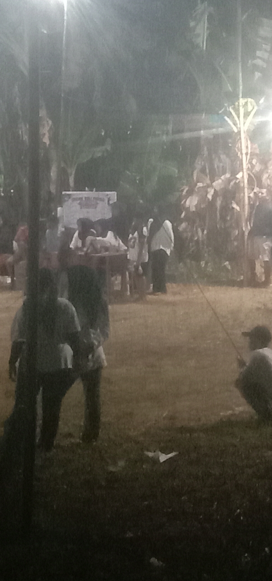 The image depicts a nighttime outdoor gathering in Banjar Dowo, Indonesia, characterized by a dimly lit, hazy atmosphere, likely due to artificial lighting and possibly fog or dust. In the background, a white banner with text is visible, suggesting an event or announcement. Several individuals are present, scattered across a grassy area with some trees and foliage. One person on the right is crouched down, holding what appears to be a fishing rod. Other people are standing or moving around, some gathered near a table or platform. The overall scene suggests a casual social event.