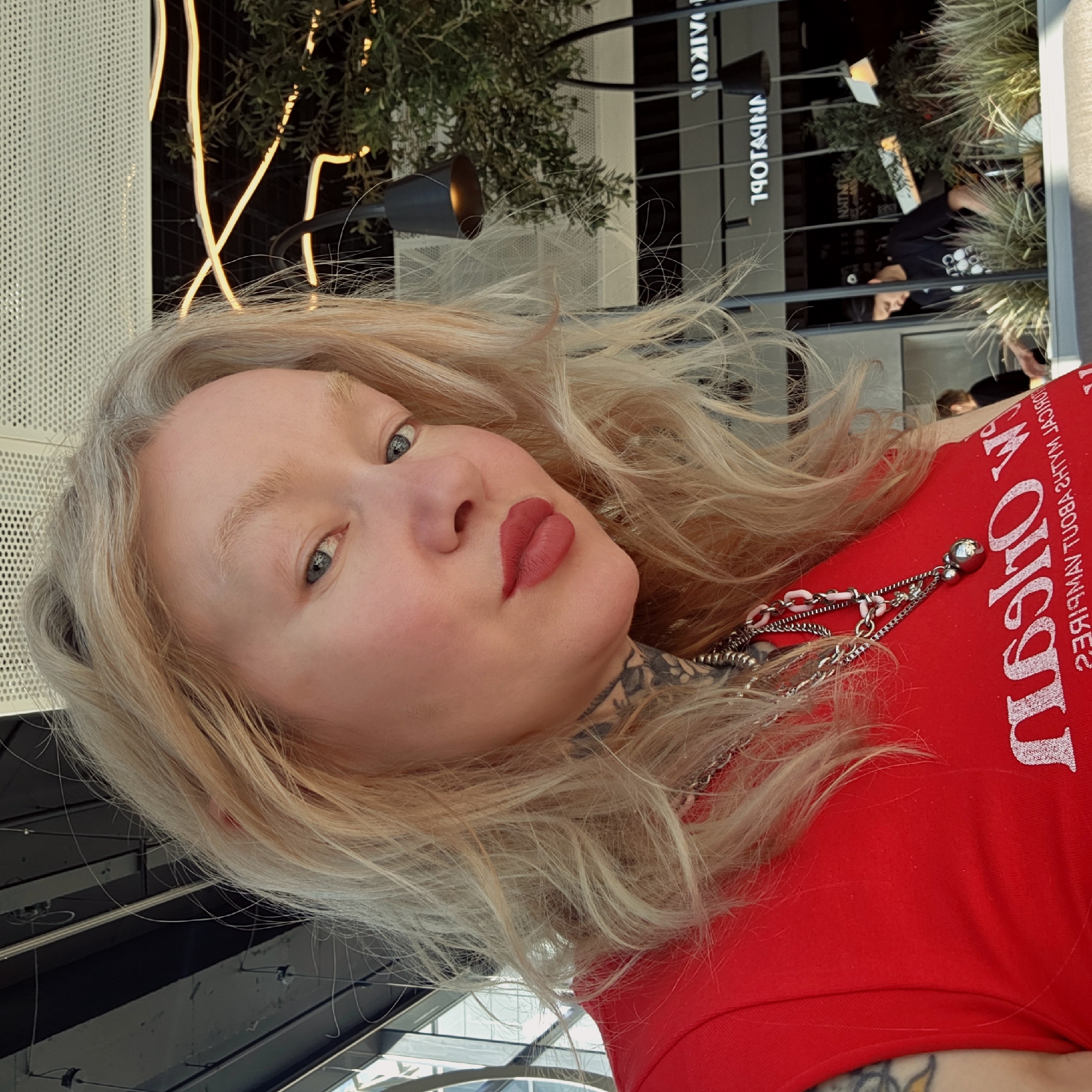 A portrait-oriented photo captures a fair-skinned person with long, wavy blonde hair and blue eyes, looking directly at the camera. The person is wearing vibrant red lipstick and a red top. A silver chain necklace with a small pink link detail and a metallic sphere charm is visible around their neck. Tattoos are visible on the person's neck and left arm.
The image is taken from a slightly high angle, suggesting a tilted or laid-back pose. The background indicates an indoor or covered outdoor setting, likely a modern cafe, restaurant, or shopping complex in Saint Petersburg, Russia.
Behind the person, the setting includes a textured light-colored wall on the left and a darker, reflective surface on the right. Artificial yellow lights with a linear design are visible, along with dark green foliage from potted plants. Reflected in the background are various architectural elements, light fixtures, and indistinct figures of other people. The time of day appears to be bright, with both natural and artificial light illuminating the scene.
Visible text includes:
* On the red shirt (mirrored): "I'M QUEEN / ABOUT THAT SHIT MYSELF"
* In the background (mirrored, possibly signage): "DAIKON" and "1907HNA" (likely part of a larger name or address).