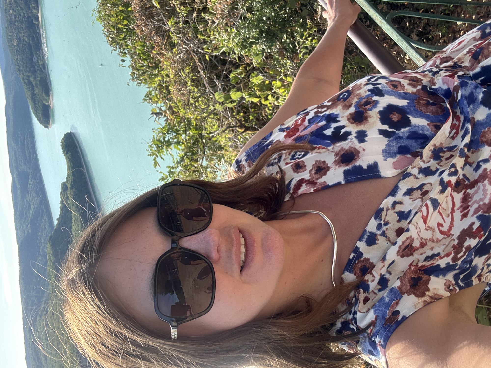 A woman with brown hair, wearing sunglasses, a silver necklace, and a floral print dress, poses for a selfie outdoors. She is looking towards the camera and smiling. The background shows a body of water, possibly a lake, surrounded by green hills. The scene is likely located at the Lac de Vouglans, a reservoir in the Jura department of France, near Moirans-en-Montagne. The sky is clear and sunny.