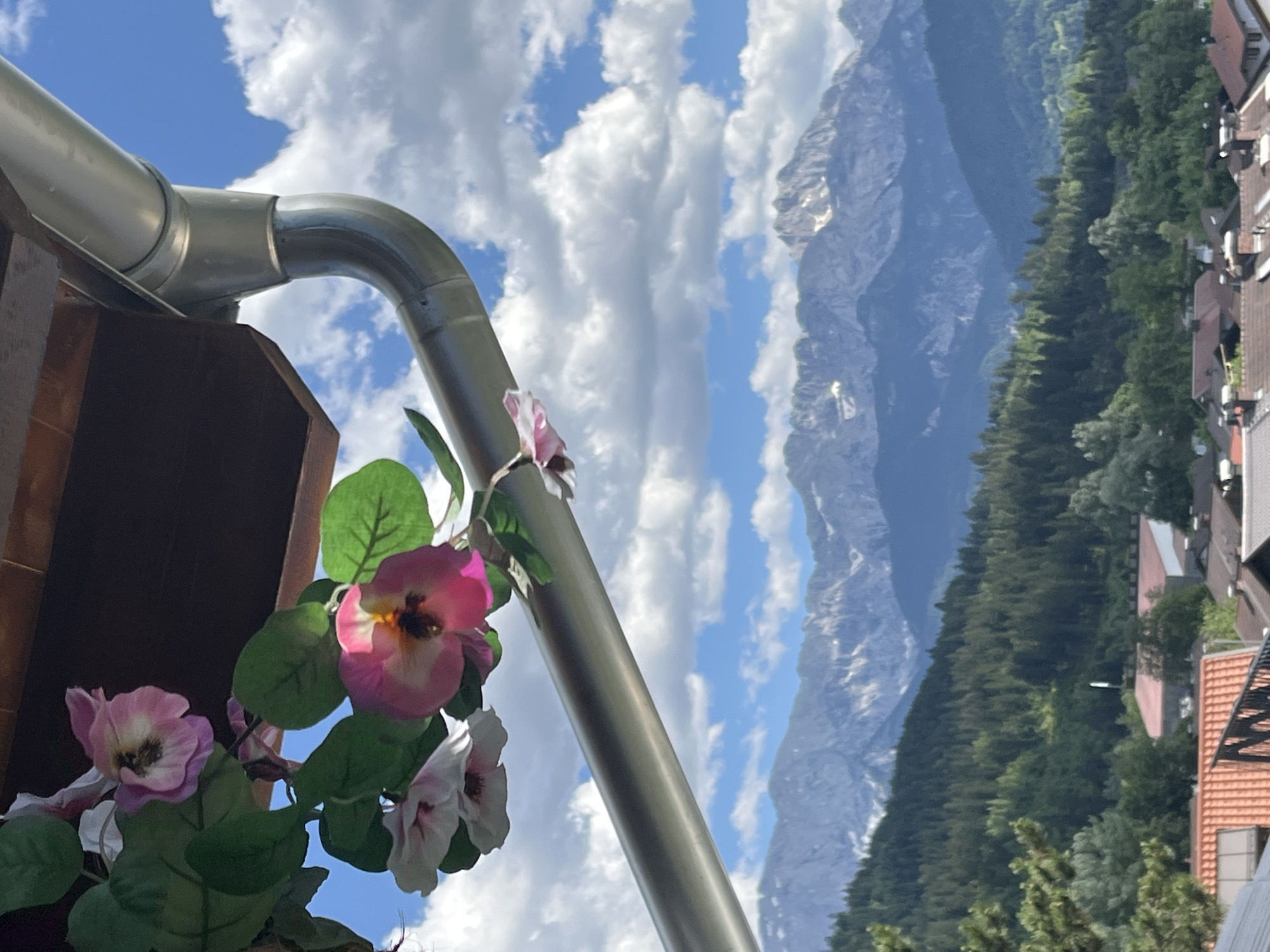 The image depicts a picturesque view of the Bavarian Alps from Garmisch-Partenkirchen, Germany. In the foreground, a metallic gutter system runs diagonally across the frame, with artificial pink and white petunia flowers hanging from the side. The gutter is attached to a wooden structure. Behind this, a lush green forested hillside slopes down towards a village with terracotta-tiled roofs. The majestic snow-capped peaks of the Alps form the backdrop, under a sky filled with fluffy white clouds.