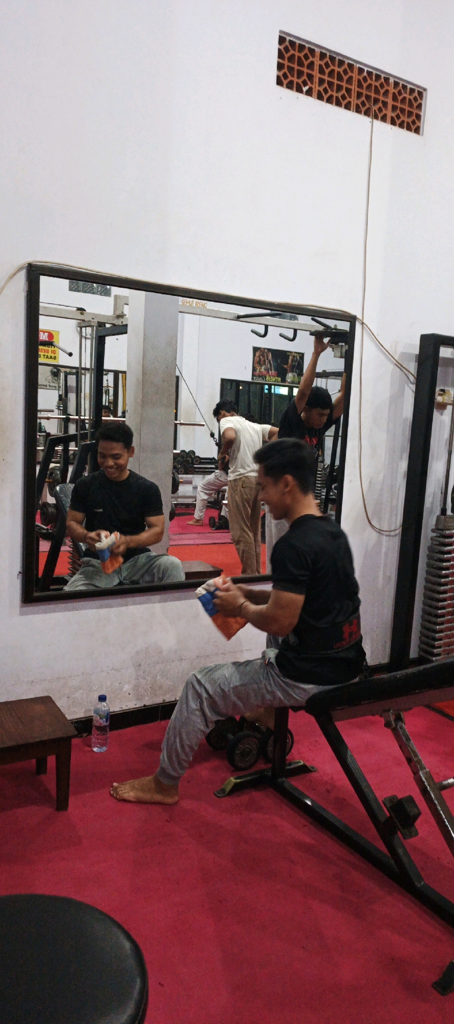 The image depicts the interior of a gym, with a large mirror reflecting a section of the room and the people within it. The gym has a vibrant red carpet floor. In the foreground, a person sits on a weight bench, wearing a black t-shirt and grey sweatpants, and holding an orange and blue towel. Their bare feet rest on the carpet. To the left of this person, a small wooden table and a bottle of water are visible.
The mirror shows three other individuals. One person, in a black t-shirt, is seen sitting and looking into the mirror, also holding a towel. Another person, in a white t-shirt and light-colored pants, is standing and appears to be stretching or performing an exercise. A third person, in a black t-shirt, is reaching up to a pull-up bar. Behind them, gym equipment, including weight racks and dumbbells, can be seen. The walls are white, and a decorative vent with an intricate pattern is visible near the ceiling. The gym appears to be located in Kasiyan Timur, Indonesia.