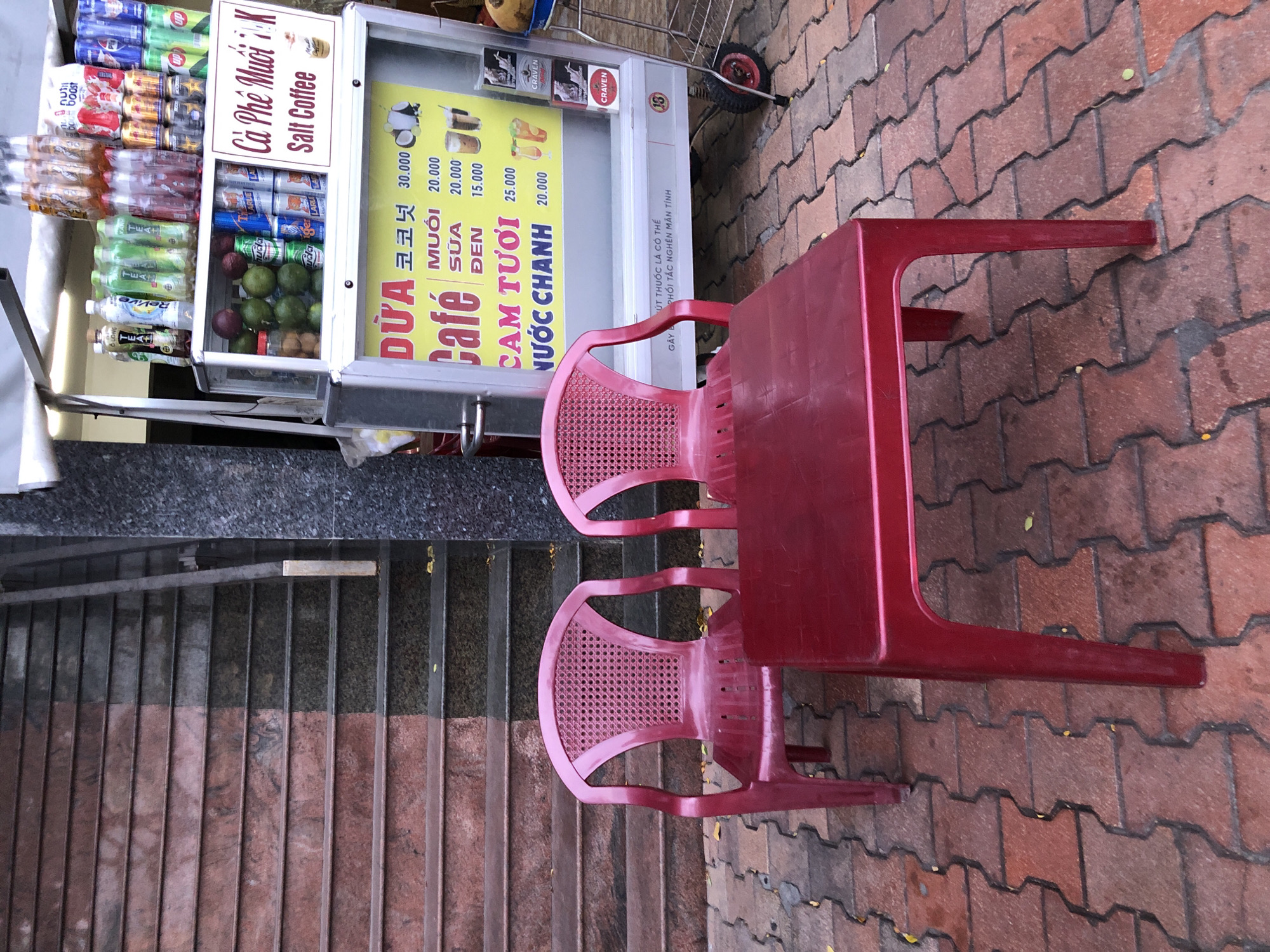 A red plastic table and two red plastic chairs are set up on a red brick walkway in front of a street food cart. The cart has a refrigerated display case filled with fruit and beverages, and an advertising sign above it that lists various drinks and prices in Vietnamese and English. The sign says "DỪA CAFE" (Coconut Coffee), "MUỐI SỮA ĐEN" (Salt Milk Black) with prices 20.000, 20.000, and 15.000, and "CAM TƯƠI NƯỚC CHANH" (Fresh Orange Lemonade) with prices 25.000 and 20.000. The cart is branded with the words "CÀ PHÊ MUỐI K" and "SALT COFFEE" along with an image of a coffee cup. Behind the cart, a section of reddish-brown granite stairs ascends to the left, and a grey granite wall with some leaf litter on the stairs is to the right. The overall scene suggests a casual outdoor seating area for a beverage vendor in the city of Đà Nẵng, Vietnam.