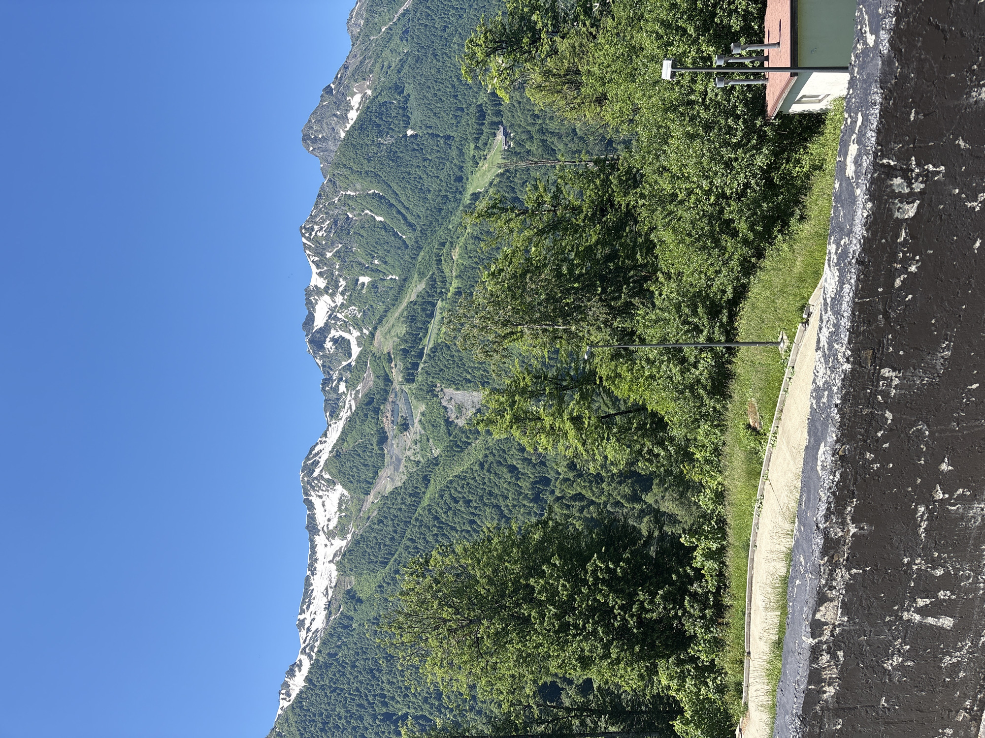 The image shows a view of the Caucasus Mountains from a location in Estosadok, Russia, on a clear, sunny day. In the foreground, a weathered, dark-colored concrete barrier runs across the bottom of the frame. Behind the barrier, a paved walkway with some grassy areas curves to the right. Further back, a dense forest of green trees covers the lower slopes of the mountains. The upper reaches of the mountains are rugged and rocky, with patches of snow still visible on the peaks and higher elevations. A ski lift track or path is visible winding down one of the mountain slopes. A small, light green building with a reddish-brown roof and a couple of pipes on top is situated among the trees on the right side of the image. A streetlamp stands next to the building, and another streetlamp is visible in the middle ground. The sky is a vibrant, cloudless blue. There are no people visible in the scene.