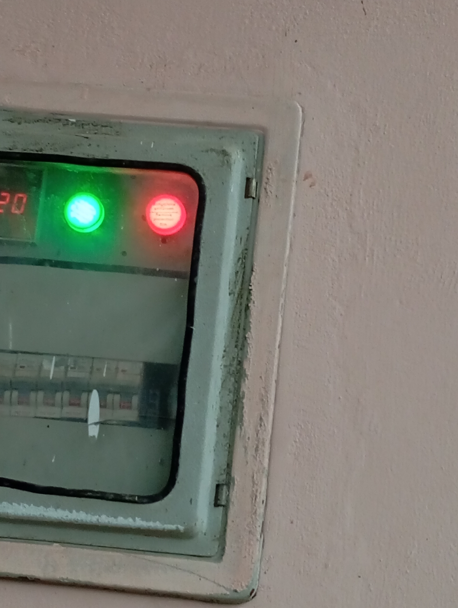 The image shows a close-up of an electrical panel mounted on a light-colored, textured wall. The panel itself is a light greenish-blue color with a weathered appearance and appears to be made of metal. It has a hinged door with two small hinges visible on the left side.
Inside the panel, the top section displays a digital readout that shows the number "20" in red digits on the left. To the right of the digits, there are two indicator lights. The left light is a bright, solid green. The right light is a red light, also solid. The surface of the red light appears to have some text or a pattern visible within it.
Below the indicator lights, the panel contains a series of what appear to be circuit breakers or switches. These are arranged in rows and have small white levers or handles. The lower portion of the panel is darker and less clear, but it seems to house these electrical components.
The overall environment appears to be indoors, possibly in a utility area or a hallway. The wall has a slightly rough texture and a light, neutral color, showing some minor smudges or discoloration in places, particularly around the edges of the electrical panel. The lighting in the scene is relatively even, highlighting the illuminated indicators on the panel.