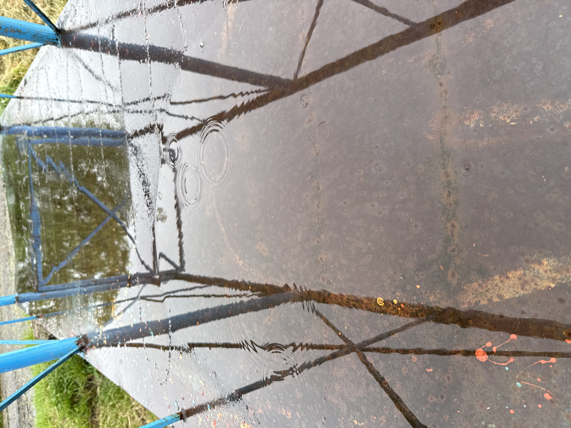 The image depicts a close-up, downward-angled view of a shallow pool of rainwater on an oxidized metal surface in Mykhailivka-Rubezhivka, Ukraine. The majority of the frame is occupied by the water, which reflects elements from the surrounding environment. Concentric ripples are visible in the upper mid-section of the water, indicative of recent raindrop impacts.
The metal surface beneath the water is visibly rusted, displaying a mottled pattern of dark brown, orange, and reddish-brown hues, along with a textured or pitted appearance. Splatters of bright orange/red and light blue paint are present on the lower left and mid-right areas of the surface.
Reflected in the water are multiple vertical and diagonal structures. Two prominent blue metal bars or posts are reflected from the upper left and upper right edges of the frame, their reflections distorted by the water's movement. Additional darker, structural reflections, possibly from an overhead frame or support, are also present, particularly in the upper right and mid-left. A patch of green vegetation, consistent with grass, is faintly visible in the reflection towards the upper left. No individuals or animate subjects are present in the scene.