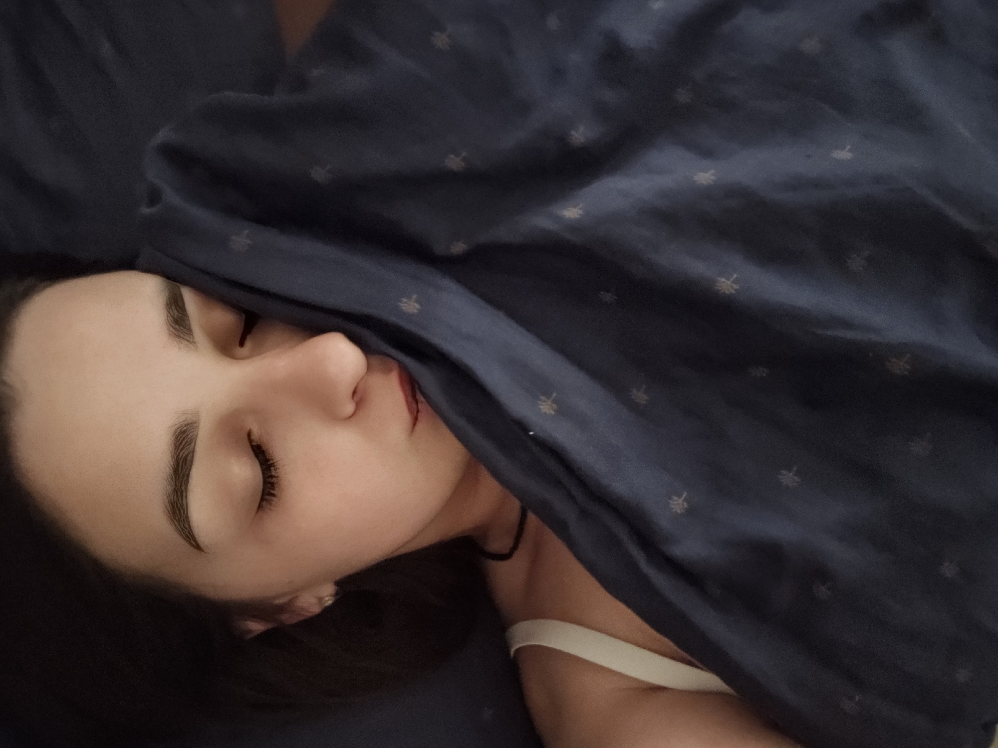 A close-up, overhead shot shows a young woman with her eyes closed, lying in bed. Her face is partially obscured by a dark blue blanket with a subtle pattern of small, light-colored symbols. The woman has dark hair, arched eyebrows, and is wearing a light-colored strap, possibly from a camisole or bra. She also has a thin, dark necklace. The scene appears to be an indoor setting, likely a bedroom. There is no information in the image to suggest the location is in Petropavlivska Borshchahivka, Ukraine.