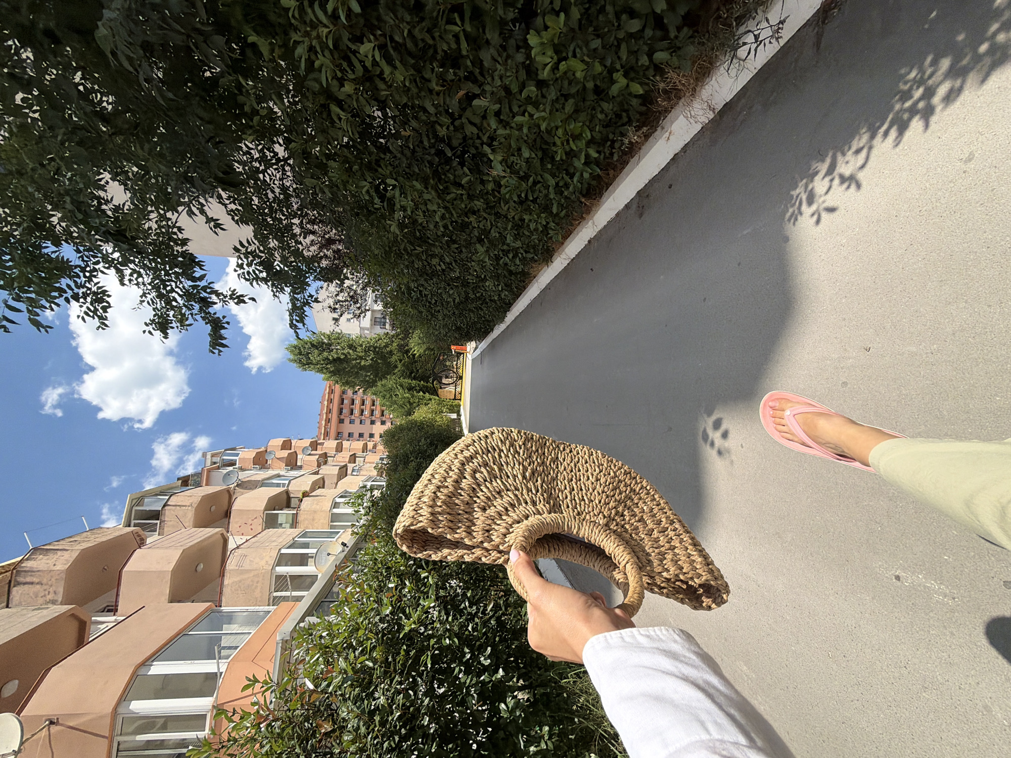 A person's right hand is holding a woven, beige bag in the foreground, with a person's left leg and pink flip-flop visible below. The scene appears to be a residential street in Budva, Montenegro, with a road extending into the distance, bordered by lush green hedges on the right and a multi-story, peach-colored building with balconies on the left. The sky is blue with scattered white clouds, and shadows of leaves are cast on the pavement.