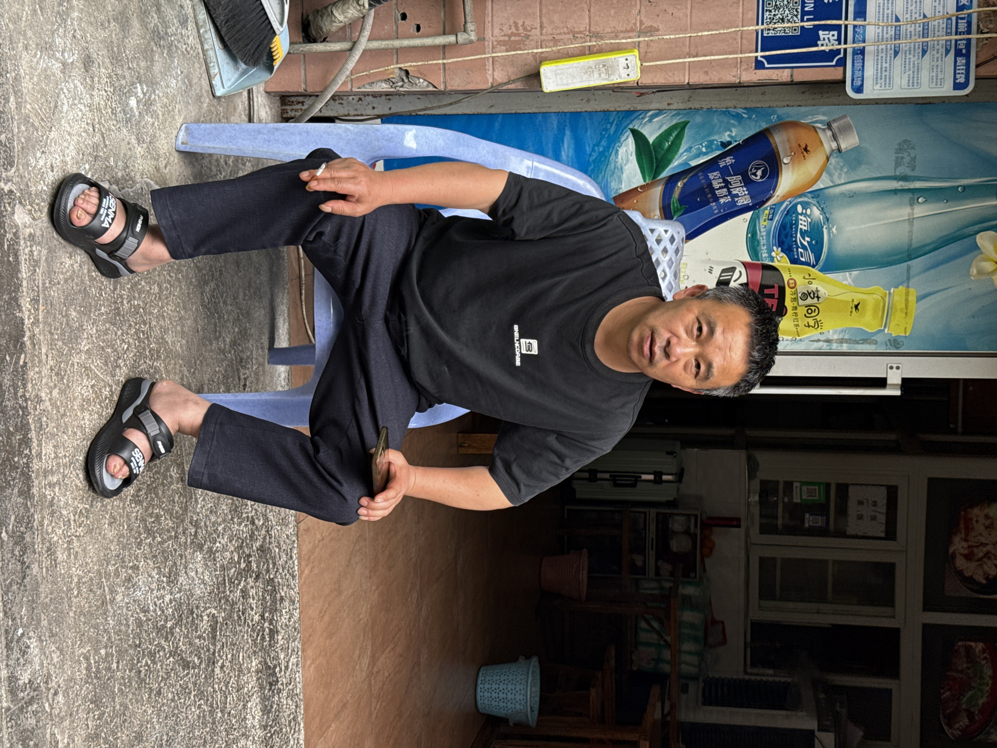 A man is sitting on a blue plastic chair. The location appears to be a street corner in the High-tech Zone of Chongqing, China. He wears a black T-shirt, dark pants, and black sandals. He holds a cell phone in his right hand. The man is looking directly at the camera. Behind him is an advertisement for beverages. The scene is partially outdoors with a cement floor and partially indoors with a tiled floor. Other objects visible include a broom, a water bucket, a suitcase, furniture, and dishes in display cases. The OCR text visible on the advertisement includes Chinese characters and phrases such as "High-tech Zone, Chongqing, China," and names of beverages such as "Sea Salt Lemon" and "Tongyi Assam."