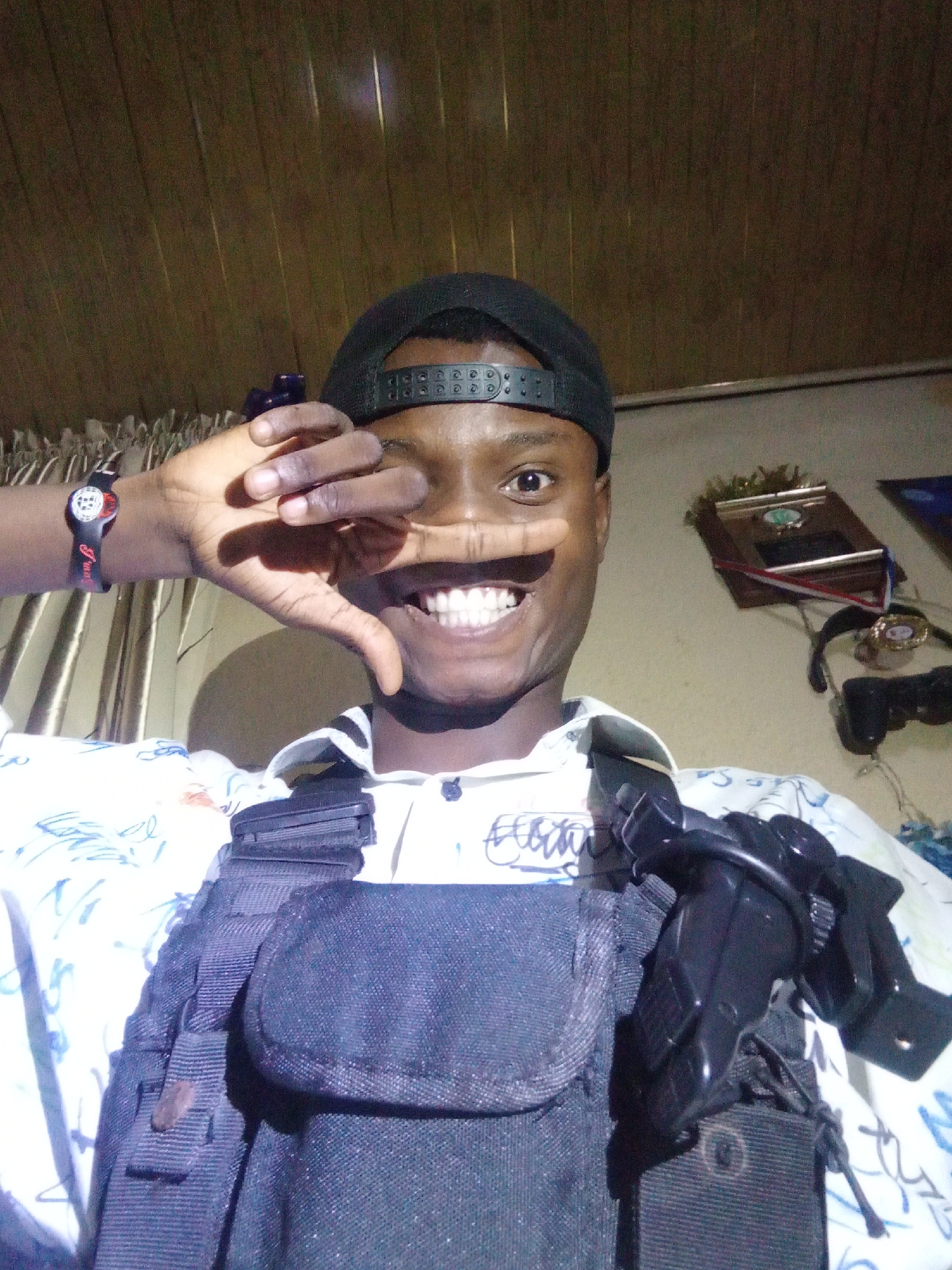 A young Black man in an indoor setting, possibly in Narayi, Nigeria, is seen from a low angle, smiling and obscuring one eye with his hand. He is wearing a black baseball cap, a white shirt with a pattern, and a dark-colored tactical vest with a visible pistol holster. Behind him, there are vertically-striped wooden panels, a framed award or plaque, and video game controllers hanging on the wall. He is wearing a wrist watch on his left hand.