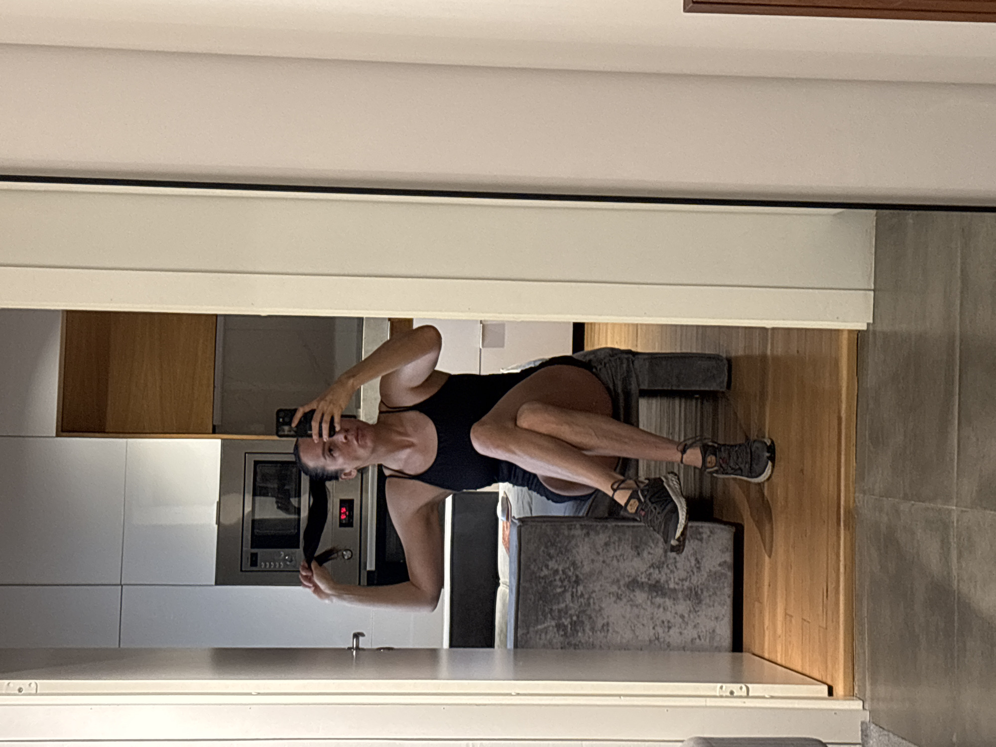 The image captures a woman taking a mirror selfie in what appears to be a modern indoor living space, likely an apartment in Chaikovsky, Russia. The woman, in her 30s or 40s, is seated on a grey upholstered surface, possibly a couch or armchair, with her legs bent. She has dark hair pulled back and is wearing a black one-piece swimsuit or athletic attire. She holds a black smartphone in her right hand to take the photo, while her left hand is near her hair. She is wearing dark athletic shoes with white soles and intricate strap details.
The reflection in the mirror reveals a contemporary kitchen area behind her. This kitchen features sleek white lower cabinets and a section of wooden-toned upper cabinets. A built-in stainless steel oven or microwave is visible, displaying the digits "09" on its digital clock or timer. The flooring transitions from light wooden planks in the foreground to what seems to be grey tiled or concrete flooring further back, with a vertical wooden panel acting as a partition. The lighting in the scene is artificial, typical of an indoor evening or night setting. The woman's expression is neutral, focused on the act of taking the picture.
