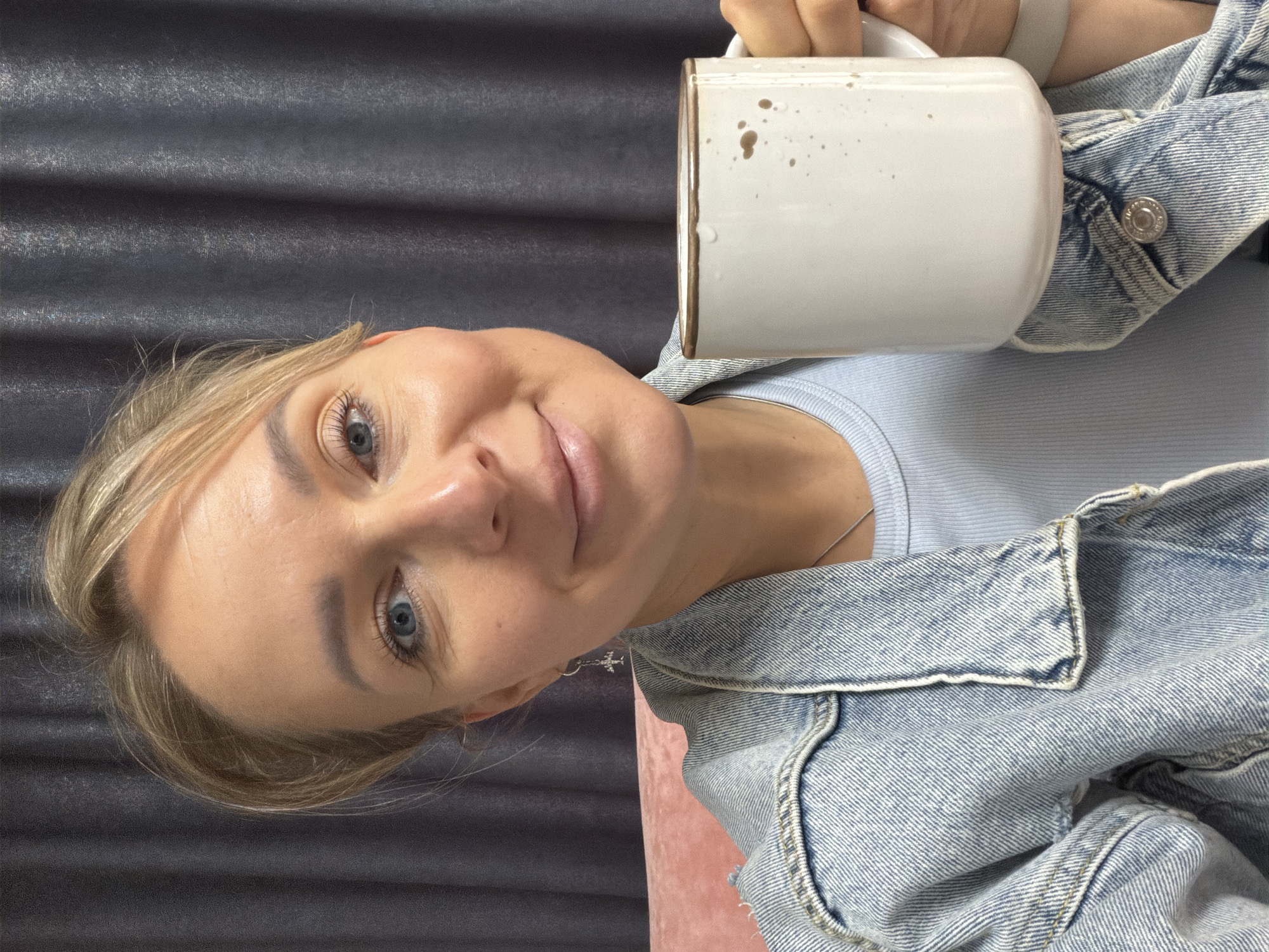 The image features a woman captured from an upward-looking perspective, cropped from the chest up, in an indoor setting within Chaikovsky, Russia. She has blonde hair, blue eyes, and fair skin, and is looking towards the viewer with a gentle, pleasant smile. Her hair is parted on the side, framing her face, and she appears to be wearing light makeup.
The woman is holding a large, off-white ceramic mug in her right hand, which is prominently positioned in the upper right of the frame. The mug has small dark speckles and a few visible brown drips or stains on its side, suggesting it contains a beverage like coffee. She is dressed in a light blue ribbed top or t-shirt, layered under a light blue denim jacket. A thin silver chain necklace is visible around her neck, and a small, cross-shaped earring can be seen on her left ear.
The background primarily consists of a dark grey or black textured fabric, possibly a curtain, with subtle horizontal ridges. A soft, pinkish, possibly velvet, surface, likely a piece of furniture, is partially visible on the right side of the background. The lighting suggests an indoor environment, possibly during the daytime or a well-lit interior. The overall scene conveys a sense of casual comfort and contentment. There is no discernible text visible on any objects or clothing.