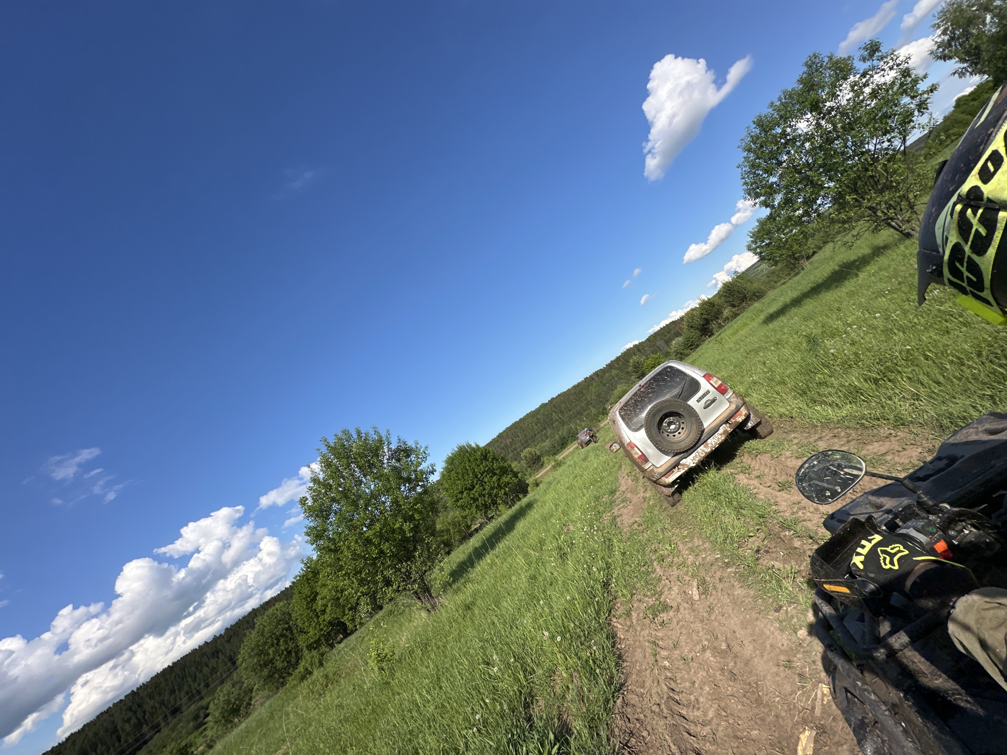 The image depicts a landscape with a dirt track running through a grassy field in the direction of a forest, possibly in the vicinity of Пепеляева, Russia. A silver SUV, with a spare tire mounted on the back, appears to be parked on the track ahead. In the foreground, part of an all-terrain vehicle (ATV) is visible, including a mirror and the rider's hand wearing a glove with "FOX" branding. The sky is blue with scattered clouds. The image is angled as if taken from the perspective of the ATV rider. The scene is outdoors, and the environment suggests an off-road or rural setting.