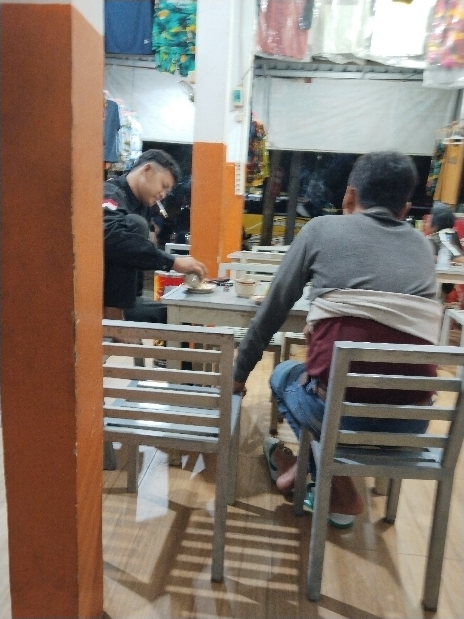 The image depicts a casual indoor scene, possibly a cafe or small eatery, with several individuals seated at tables. In the foreground on the left, a man wearing a black jacket with red and white accents is seated, actively smoking a cigarette and holding a small cup and saucer. He appears to be looking down at the table. To the right, another man, seen from the back, is wearing a grey long-sleeved shirt and blue jeans. A beige cloth is tied around his waist. He is seated and facing away from the camera, looking towards the same table area. Other patrons are visible in the background, including a person wearing a red shirt. The environment features simple tables and chairs, with what appears to be merchandise displayed on shelves or racks in the background. The overall lighting suggests an indoor setting, possibly during evening hours. The scene is situated within a commercial establishment in Prigi, Indonesia.