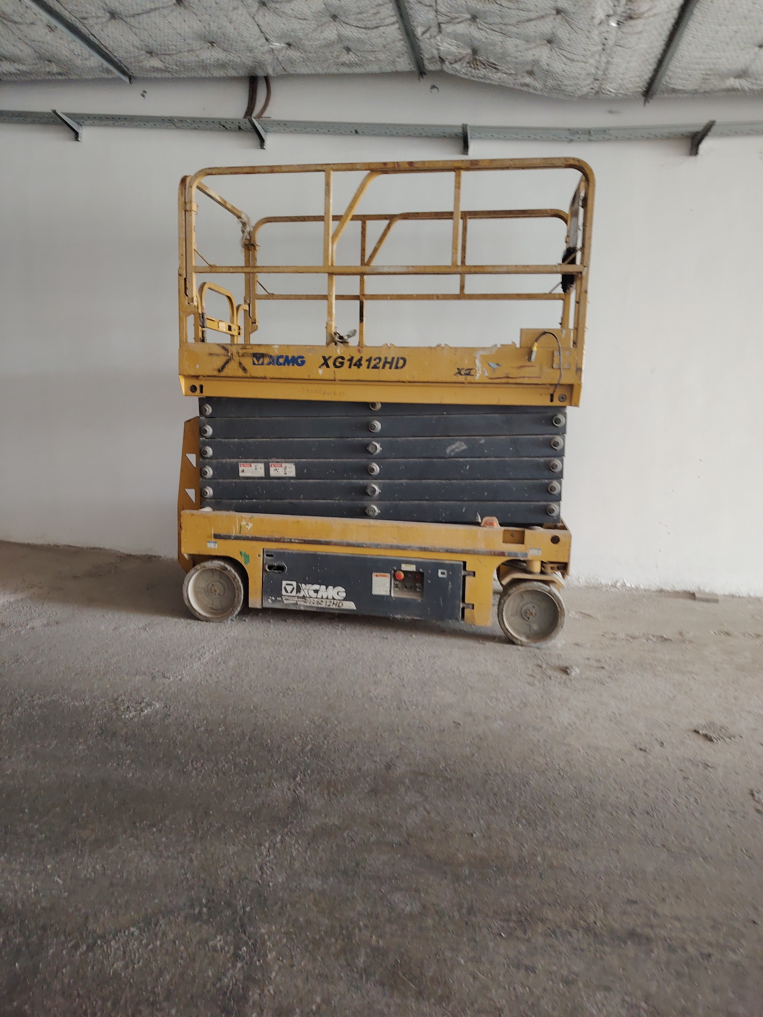 A yellow XCMG XG1412HD scissor lift is positioned in the center of the frame against a white wall in an interior space. The scissor lift has a grey platform with several stacked horizontal panels. The lower portion of the scissor lift is also grey, with two visible wheels. The floor is a rough, grey concrete surface. Above the scissor lift, a white ceiling with exposed metal conduits and insulation is visible. The scene appears to be in an unfinished or industrial building.
