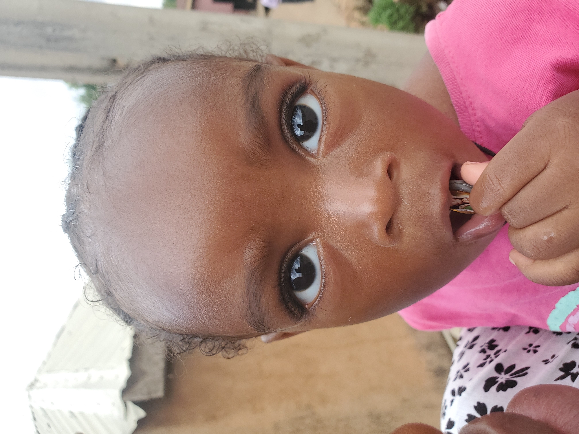This is a close-up, vertically oriented photograph of a young child. The child has dark skin and is wearing a bright pink t-shirt and a patterned skirt with white and black floral designs. The child's face is prominent, with large, dark eyes looking directly at the camera. The child's right hand is near their mouth, holding something small and brown, which they appear to be putting into their mouth. The background is blurred, but suggests an outdoor setting with some structures and greenery. The lighting appears to be natural, indicating it is daytime. The overall impression is one of candid observation, capturing a moment of childhood exploration. The image was taken in Jos, Nigeria.