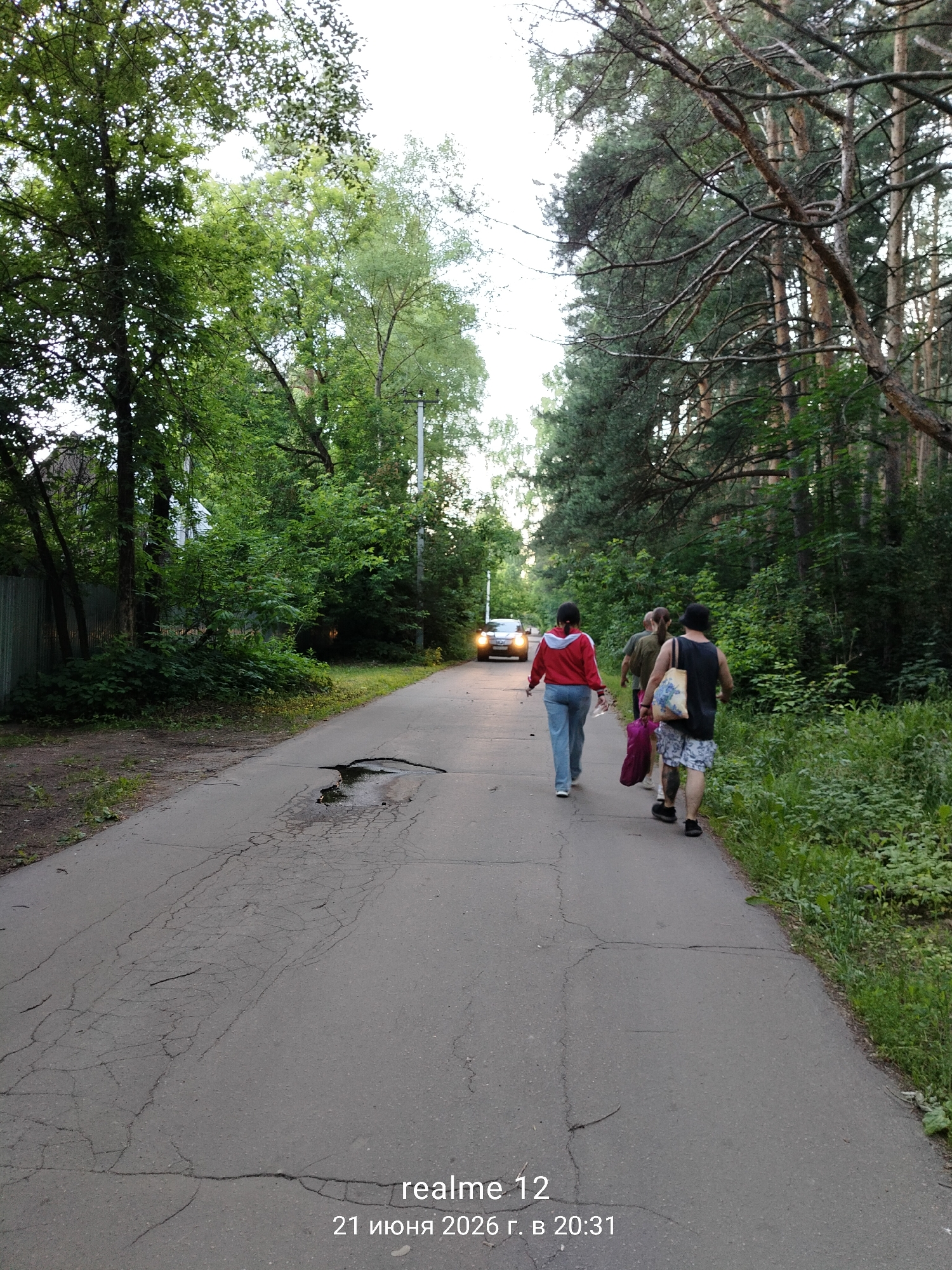 A group of three people walk down a paved road in a wooded area. The person in front, wearing a red jacket and blue jeans, walks away from the camera. Behind them, two individuals walk side-by-side. The person on the left carries a pink bag, and the person on the right wears a black tank top and patterned shorts, carrying a large tote bag. A car with its headlights on is visible in the distance, approaching the group. The road is cracked and appears to have a pothole filled with water. Lush green trees line both sides of the road, and a fence is visible on the left. The timestamp in the lower right corner indicates the image was taken on June 21, 2026, at 20:31. The location is identified as Druzhba SNT, Russia.