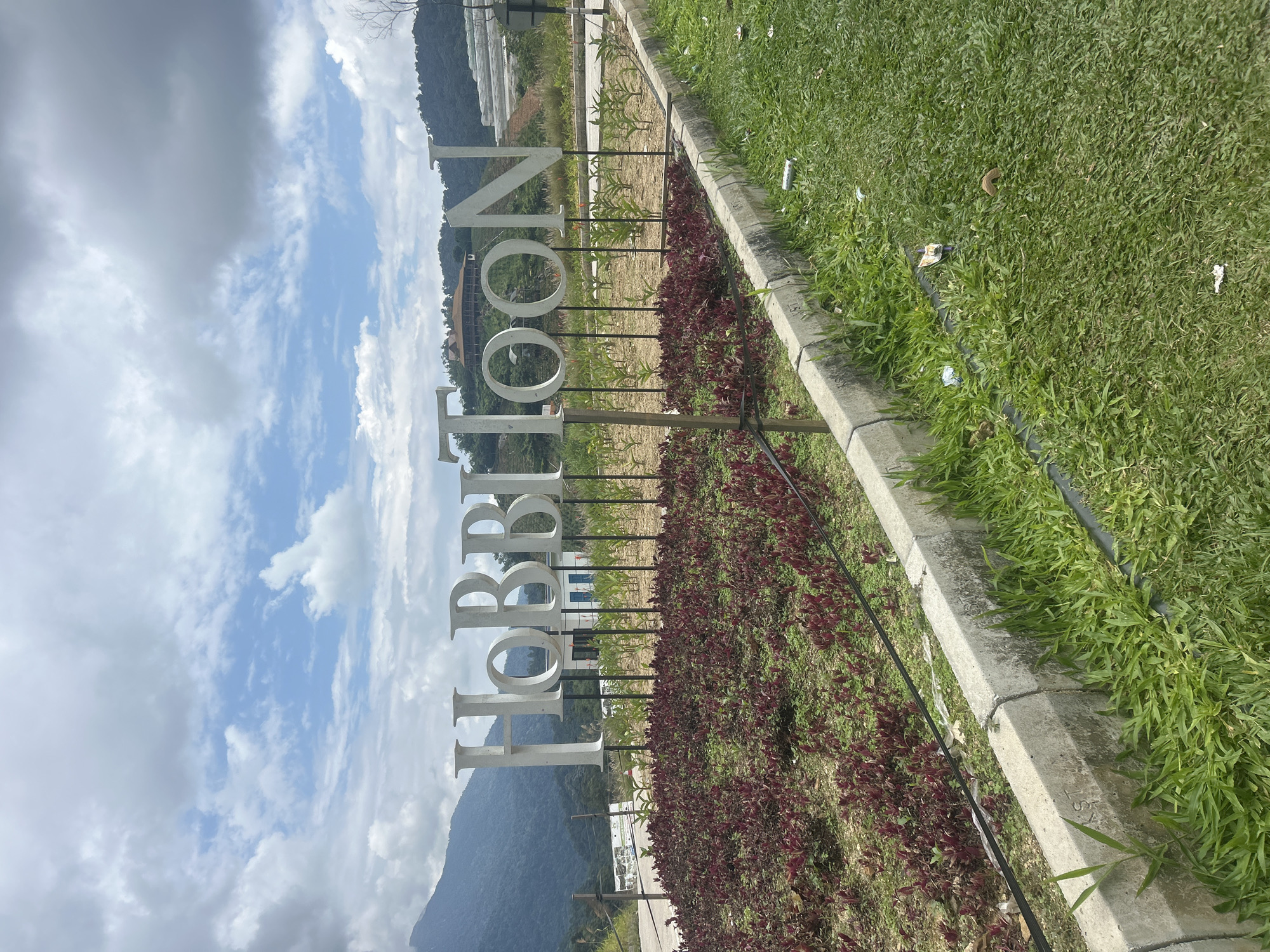 The image depicts a large, white, three-dimensional sign spelling out "HOBBITOON" in uppercase letters. The sign is positioned in an outdoor setting with lush green grass and a border of red-leafed plants in the foreground. Behind the sign, a building with a distinctive roofline is visible on a hillside, surrounded by trees and mountains. The sky above is partly cloudy with a mix of blue sky and white clouds. In the background, there appears to be some sort of terraced agricultural or horticultural development on the slopes of the hills. The overall scene suggests a tourist attraction or themed park. The location is identified as Kampung Kerawat, Malaysia. There are no people visible in the image. The scene is clean except for a few pieces of litter on the grass.