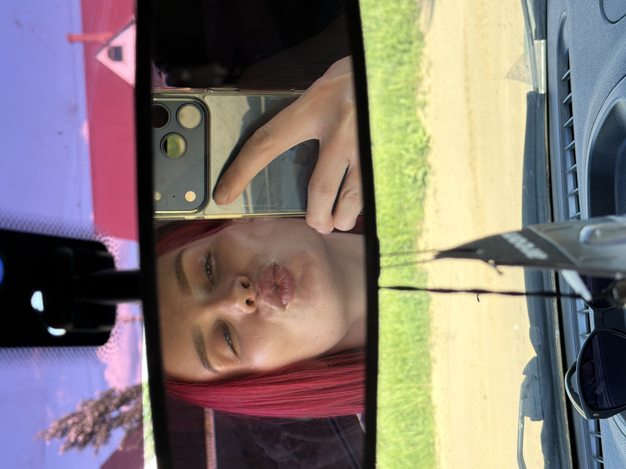 The image shows a reflection in a car's rearview mirror. A person with red hair is visible, holding a smartphone with a multi-lens camera to take a selfie. The view through the car window shows a road and grass, suggesting a rural or suburban environment. Sunglasses are visible on the right. The precise location in Chaikovsky, Russia is not determinable from the information in the image.