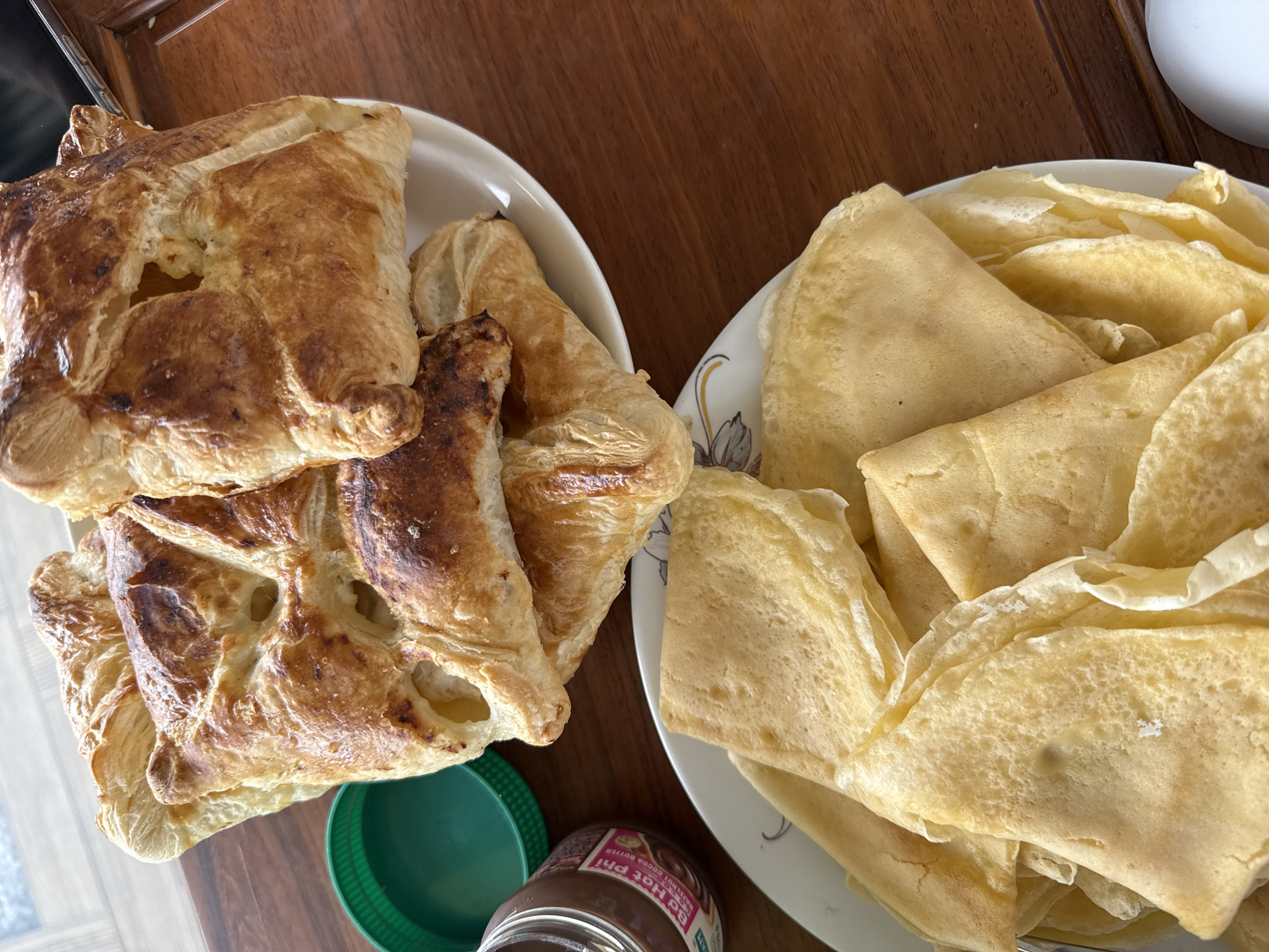 A close-up, overhead shot displays two plates of food on a wooden table. The top plate contains several golden-brown pastries, possibly croissants or turnovers, with visible layers and some browned edges. Next to this plate, a green plastic lid and a jar of chocolate hazelnut spread are visible. The bottom plate is filled with folded crepes, also golden yellow in color. The setting appears to be a casual meal, likely breakfast or a snack, in an indoor environment. The food is the primary focus, with no people or discernible activities beyond the presentation of the food. The scene lacks specific architectural details that would allow for definitive identification of a location within Bac Nha Trang Ward, Vietnam.