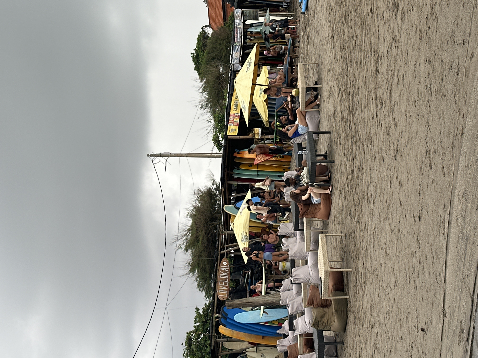An outdoor scene captures a busy beachside area, likely a surf rental or relaxation spot, under an overcast sky. The image is oriented sideways, with the cloudy sky occupying the left side of the frame and a sandy beach on the right.
In the mid-ground, a long, low structure, resembling a beach shack or surf shop, runs horizontally across the image. Numerous colorful surfboards, in shades of blue, yellow, and orange, are stacked vertically against and around this structure. Several large yellow umbrellas provide shade over groups of people seated at tables or lounging on chairs and beanbags on the sand.
Many individuals are present, relaxing and socializing. Some are seated under the umbrellas, others are standing near the surfboards, and more people are spread out across the sandy area on various seating arrangements. They appear to be tourists or locals enjoying a day at the beach, perhaps before or after surfing.
Notable details include a utility pole with wires extending from it, positioned towards the left-center of the image. The sand shows ripples and textures from foot traffic. The sky is uniformly gray and cloudy, indicating overcast weather conditions.
Visible text includes a prominent wooden sign hanging from the shack that reads "LOVELOKAL." Under one of the yellow umbrellas, a banner is partially visible, displaying the words "POSTER CANGGU SURF," confirming the location is a surf-related area, consistent with the North Kuta, Indonesia context.