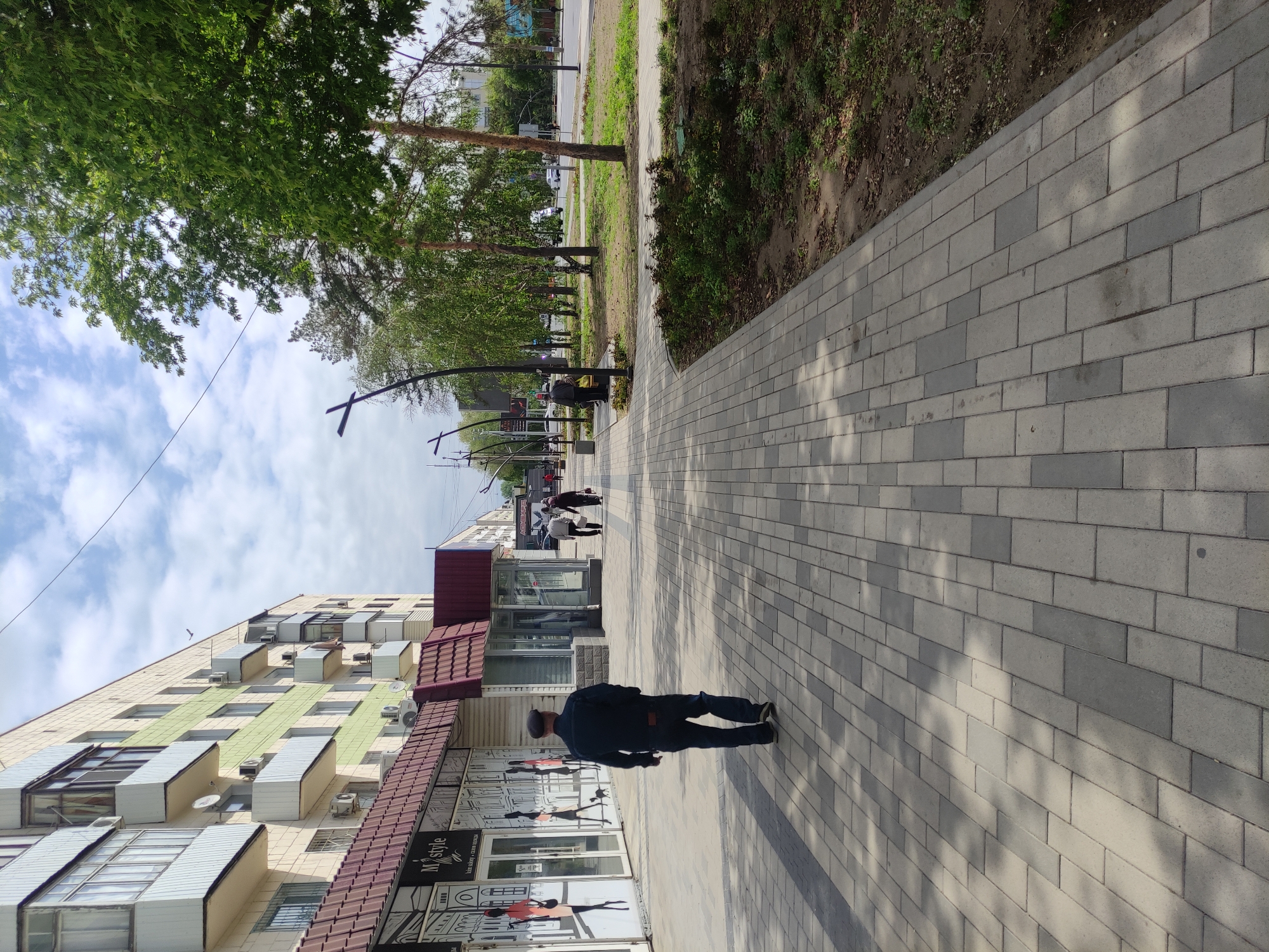 The image shows a sidewalk in Pavlodar, Kazakhstan. The sidewalk is paved with grey rectangular stones. On the left is a multi-story building with a red tiled roof and green and beige panels on the facade. A person wearing dark clothing and a cap is walking along the sidewalk towards the perspective vanishing point. Several other individuals are walking further down the sidewalk in the same direction. Trees line the left side of the sidewalk, and a grassy area is visible on the right. Light poles are positioned along the sidewalk. The sky is visible with scattered clouds. Shadows from the trees are cast on the sidewalk.