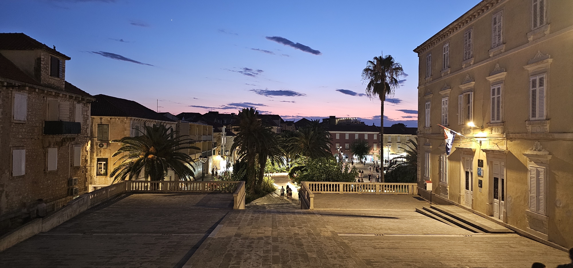 This media file captures a twilight scene in Grad Supetar, Croatia, likely during a pleasant evening. The sky is a gradient of deep blue at the top, transitioning to hues of pink and purple near the horizon, indicating dusk. A few wispy clouds are scattered across the sky.
The foreground features a paved plaza with intricate stone patterns. A low, ornate stone balustrade runs along the edge of an elevated area, overlooking a bustling square below. The architecture is characteristic of a European coastal town, with buildings constructed from aged stone and painted in warm, earthy tones, some with red-tiled roofs.
In the middle ground, a lively town square is visible, illuminated by warm artificial lights that cast long shadows. Several people are seen strolling through the square, suggesting a leisurely evening activity. Lush palm trees and other greenery are strategically placed, adding a tropical element to the Mediterranean setting. A tall palm tree stands prominently in the center of the scene.
On the right side of the frame, a building with a light yellow facade dominates. It features multiple windows with white shutters and decorative stone accents above and below each window. A Croatian flag is displayed on the facade, near an entrance with two doors. Subtle lighting highlights the building's architectural details.
On the left, another stone building with shutters and windows is visible. A dark awning or balcony structure is present on one of the upper floors. The overall mood is serene and inviting, capturing the peaceful ambiance of a European town at twilight. No visible text is discernable in the image.