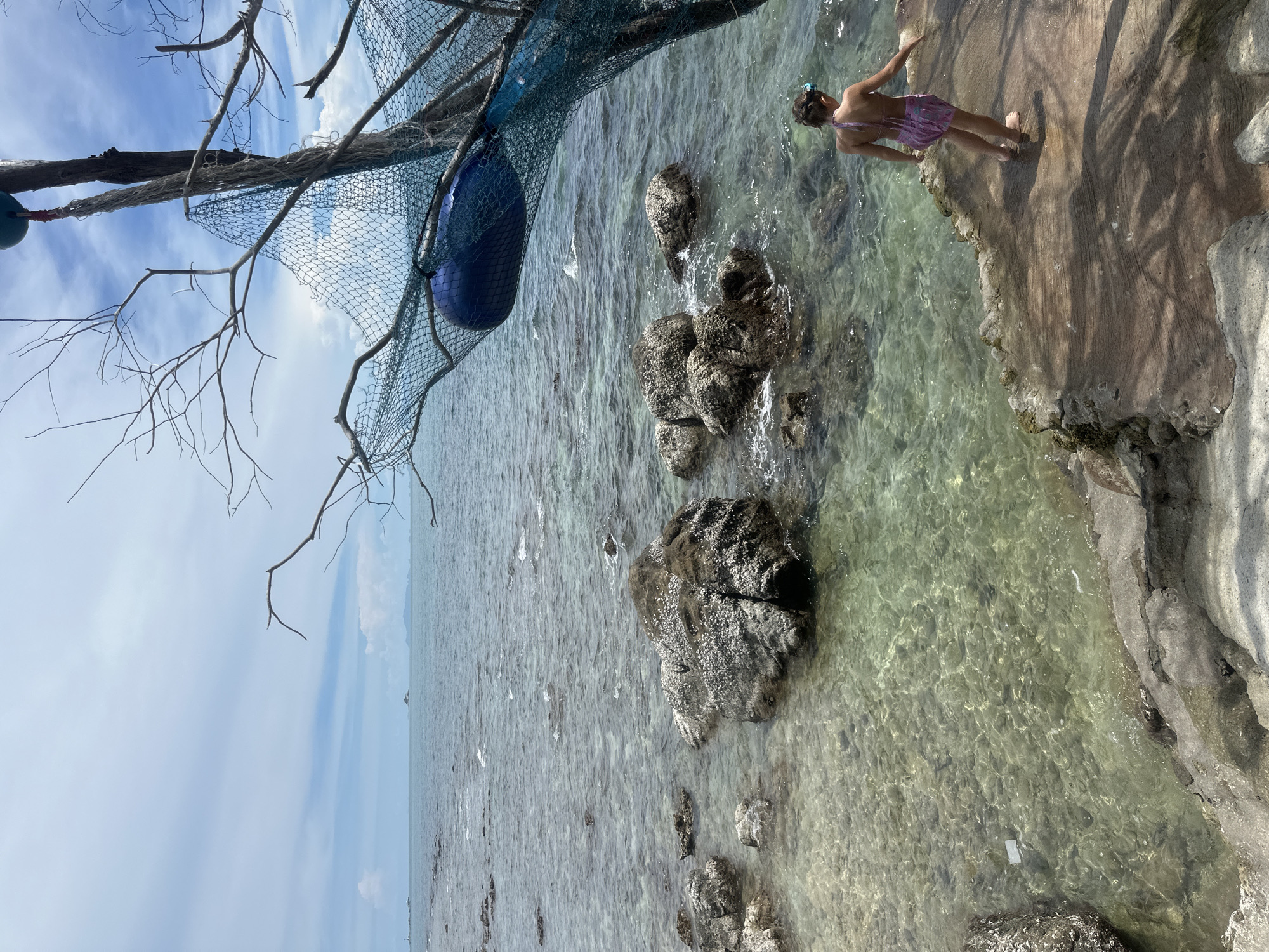 A child wearing a purple and pink swimsuit and goggles stands on a rocky shore overlooking a clear, shallow sea. The water is transparent enough to reveal the rocky seabed and scattered small rocks. In the distance, the ocean stretches to the horizon under a cloudy sky. To the right of the image, a weathered tree trunk is adorned with a blue net, a red rope, and a blue float. The child is facing away from the camera, looking towards the water. The location is identified as Ko Phangan Subdistrict, Thailand.