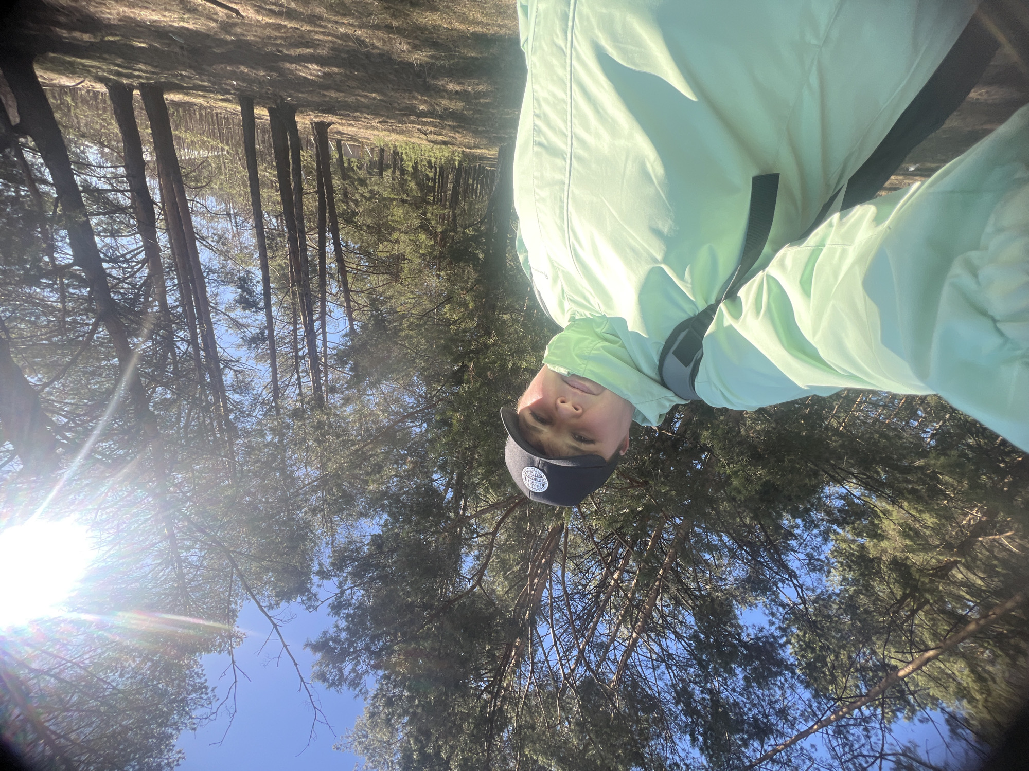A person wearing a light green jacket and a dark baseball cap is pictured in an outdoor setting with tall trees and a blue sky. The location is possibly a forest or wooded area. The photo appears to be taken at an unusual angle, with the camera tilted upward, framing the subject against the backdrop of the trees and sky. There are no discernible details to confirm the location as the city of Chaikovsky, Russia.