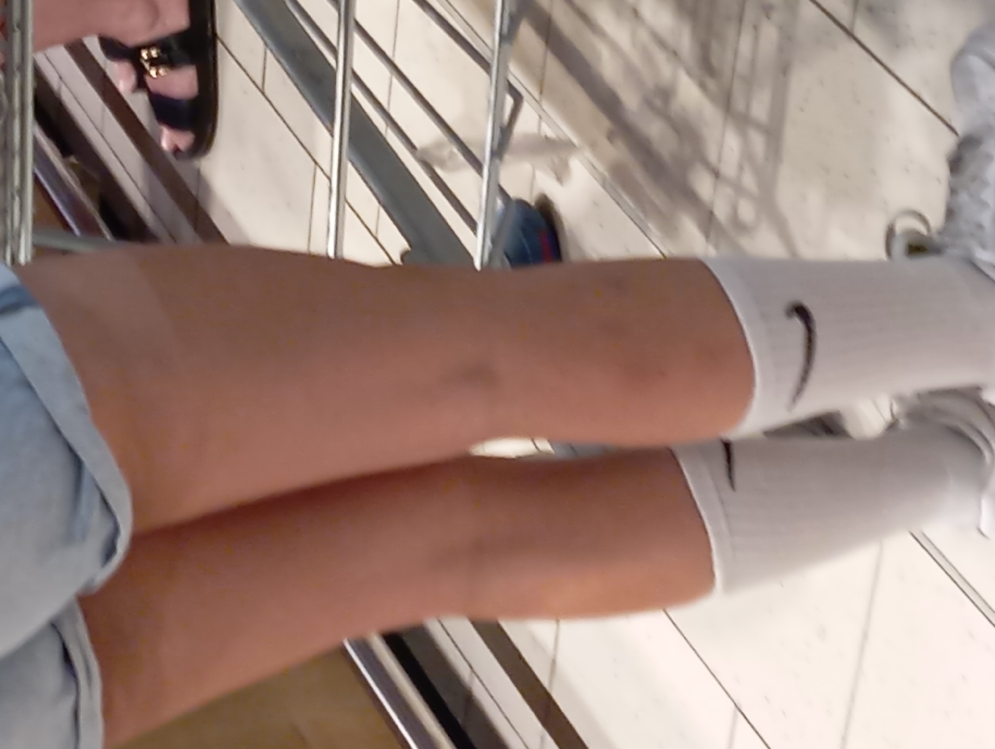 The image shows a close-up view of a person's legs and feet. The person is wearing denim shorts, white socks with black Nike swoosh logos, and white athletic shoes. The background is a tiled floor and metal railings, suggesting an indoor public space like a shopping mall or a public transit station. The lighting is somewhat dim, casting shadows on the floor. The image is taken from a low angle, focusing on the lower body. No specific landmarks in Nitra, Slovakia are visible.