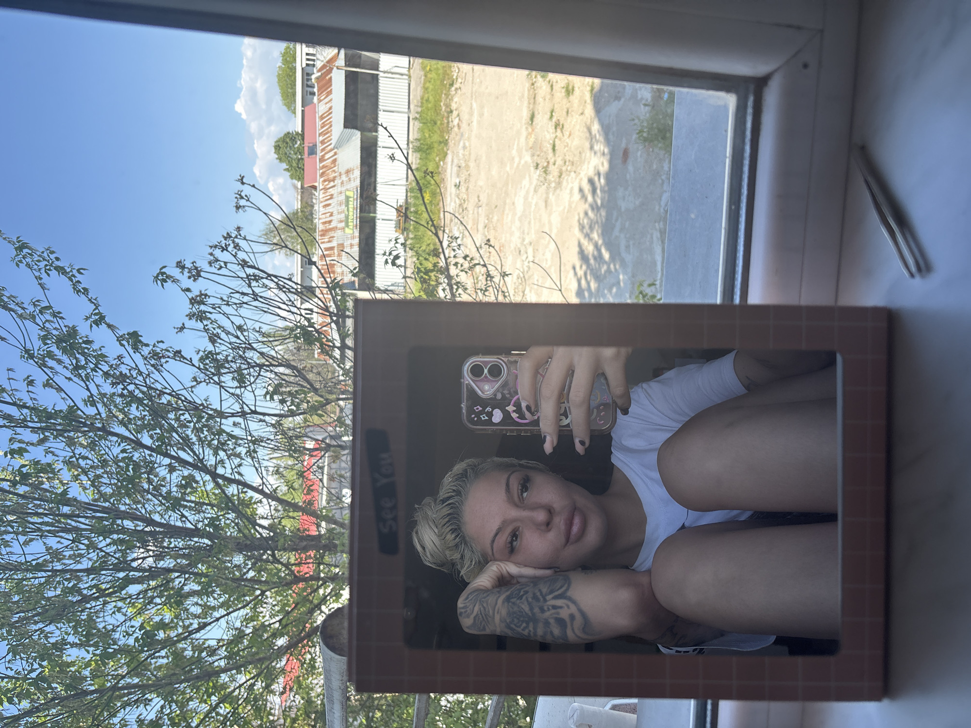 A woman is taking a selfie in a mirror, positioned in front of a window. The background visible through the window includes an outdoor scene with trees and a building. The location is in Plovdiv, Bulgaria. A pair of tweezers is present on the windowsill. The woman is wearing a white shirt and has tattoos on her arm. Her hair is blonde. She appears to be resting her head on her knees.