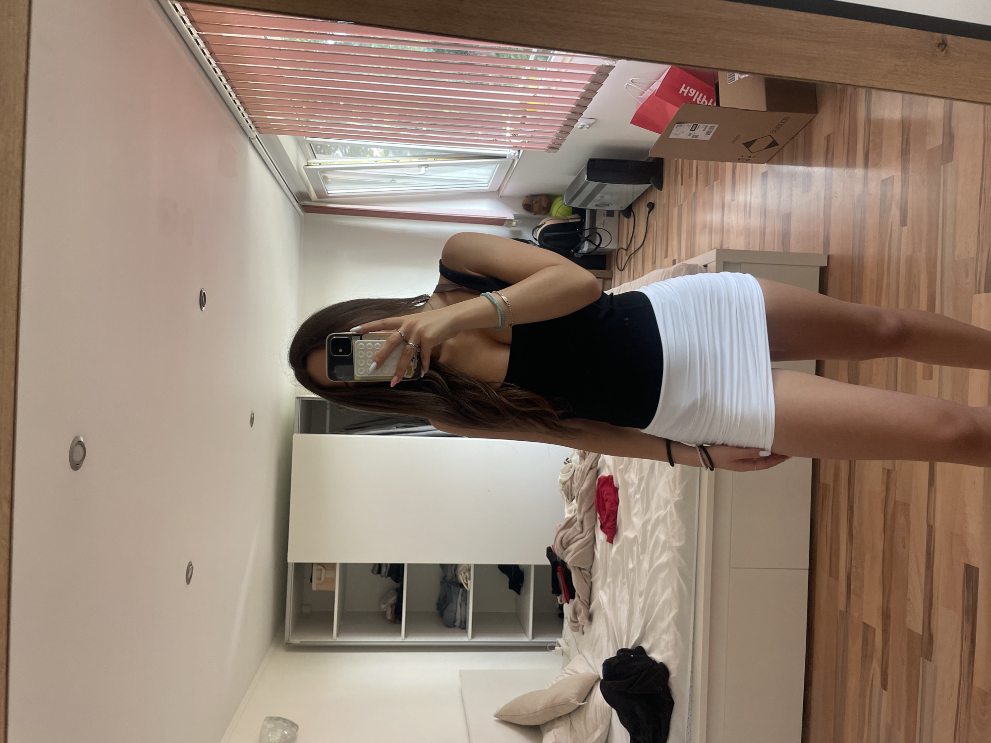 A young woman captures a mirror selfie in what appears to be a bedroom or dressing room in Varna, Bulgaria. The image is taken from a slightly low angle, showing her reflection from the mid-thigh up. She has long, dark brown hair and is wearing a black tank top and a white, pleated mini-skirt. Her left arm is raised, holding a smartphone with a white, grid-patterned case, obscuring her face. Bracelets adorn her wrists, and rings are visible on her fingers.
The room, reflected in the mirror, features a light wooden floor and white walls. A large window with horizontal wooden blinds is visible at the top, revealing bright daylight and green foliage outside. Below the window, on the floor, are several cardboard boxes, one containing a red "H&M" shopping bag, and another showing partially visible text "DO NOT EXCEE...". An electronic device, possibly a printer or speaker, sits next to them.
To the right of the woman, a white bed is visible with rumpled white bedding and pillows, along with some scattered clothes, including red and black garments. A white wardrobe or storage unit with open shelves, containing folded clothes, stands behind the bed. Recessed light fixtures are visible on the ceiling. The overall impression is a casual, lived-in private space.