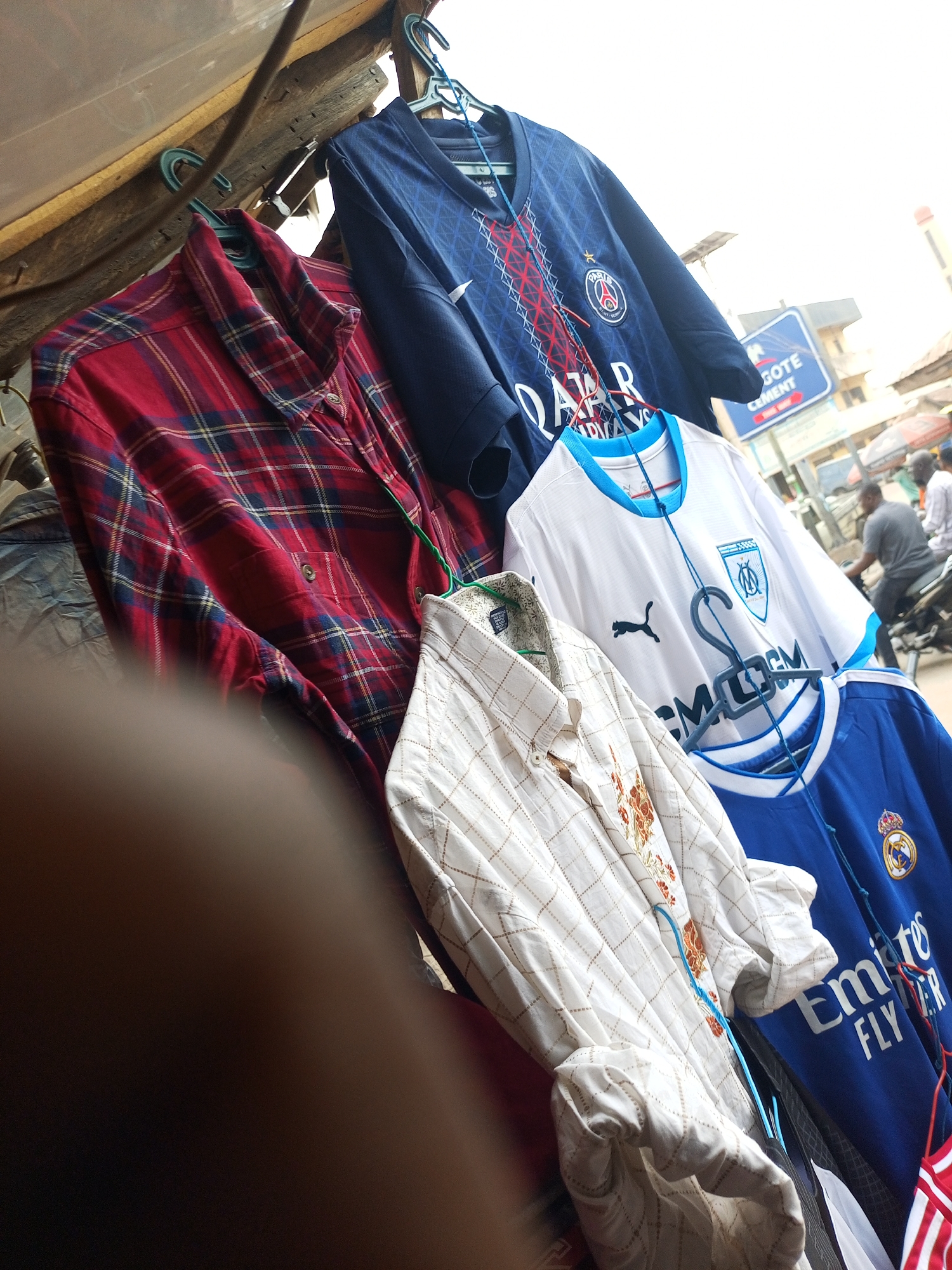 The image captures a roadside scene in Maiburiji, Nigeria, featuring a makeshift clothing rack displaying a variety of garments for sale. The rack holds shirts on hangers, including a red plaid shirt, a navy blue Paris Saint-Germain football jersey, a white Olympique Marseille football jersey, a white patterned button-down shirt, and a blue Real Madrid football jersey. In the background, there is a partially visible street scene with buildings, street signs (one reads "Dangote Cement"), and a person riding a motorbike. The lighting is bright, suggesting daytime, and the overall composition conveys a sense of a bustling, informal market setting.
