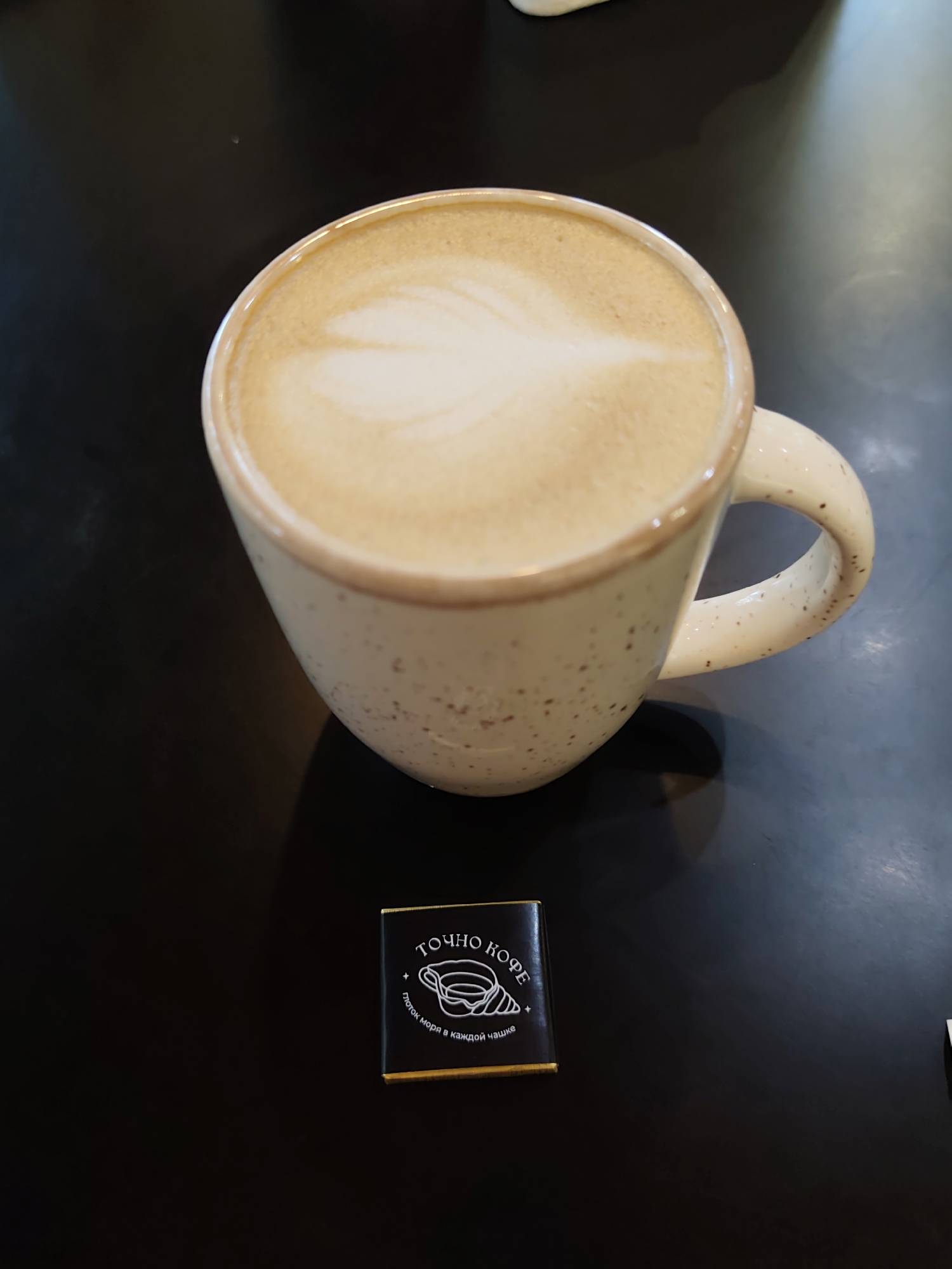 A latte in a speckled beige mug with a latte art leaf design sits on a dark, reflective surface. To the lower right of the mug, a small, dark wrapper with gold trim displays a white logo of a croissant and text in Cyrillic. The scene appears to be indoors, possibly at a cafe. There are no people visible in the image. The image does not contain any information that would indicate its location in Sochi, Russia.