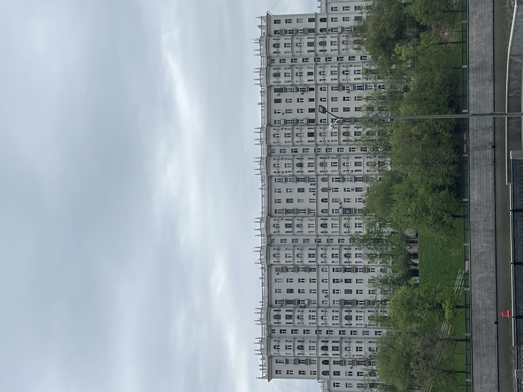 The image shows a large, white, multi-story residential building in Moscow, Russia. The building has a repeating pattern of windows and decorative architectural elements. Trees and a grassy area separate the building from a waterfront with a concrete embankment. The sky is overcast. A lamppost is visible in the foreground, and there is a red buoy on the water's edge.