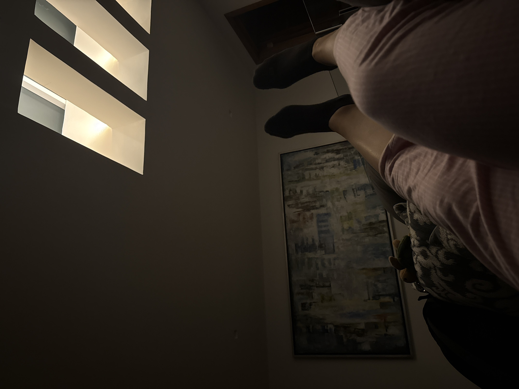 The image presents an indoor scene with dim, ambient lighting, suggesting evening or night. The perspective is from a low angle, looking upwards and across a room with dark-colored walls.
In the foreground, a person is depicted in a state of relaxation, reclining with their legs elevated and bent. They are wearing a light-colored, possibly pink, striped garment over their lap and upper legs. Their lower legs are bare, and their feet are clad in dark socks. A patterned cushion or pillow is partially visible next to them.
To the left, a striking architectural feature dominates: a section of the wall contains three vertically stacked, rectangular openings that emit a soft, warm light, creating a modern design element. On the right wall, an abstract painting with a muted palette of blues, grays, and browns hangs framed. Above the person's feet, a dark, rectangular opening, possibly a ceiling vent or a loft access, is visible. The overall atmosphere is tranquil and domestic, consistent with a contemporary living space in San Bernardino Tlaxcalancingo, Mexico. No visible text is present in the image.