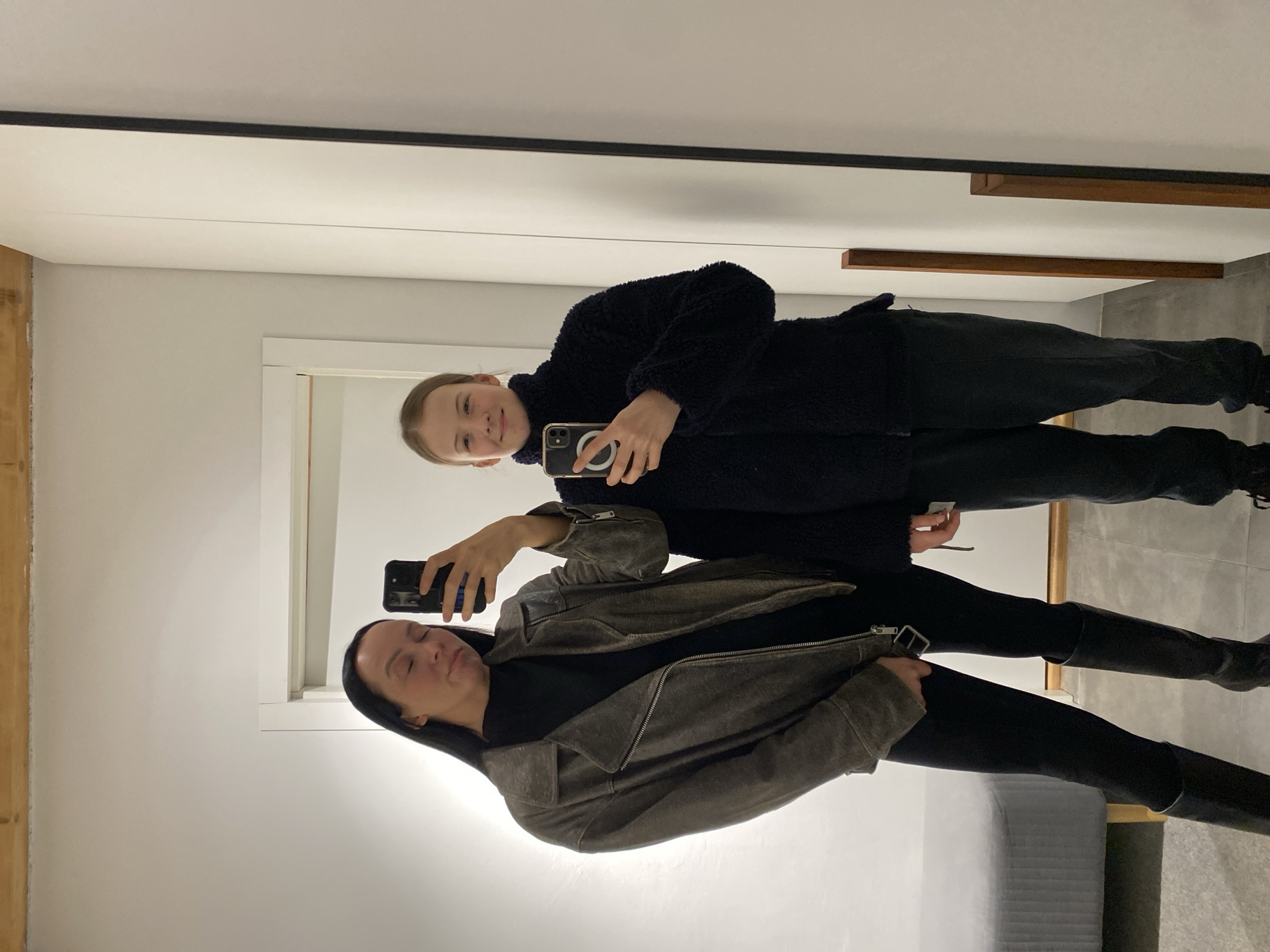 Two women are taking a selfie in front of a mirror. The woman in the foreground is wearing a gray jacket, black shirt, black pants, and black boots. She is holding a smartphone in her right hand and has dark hair. The woman behind her is wearing a dark blue fleece jacket and black pants. She is also holding a smartphone. The background appears to be a room with a door frame and a gray wall. It's not possible to confirm that this location is Chaikovsky, Russia.