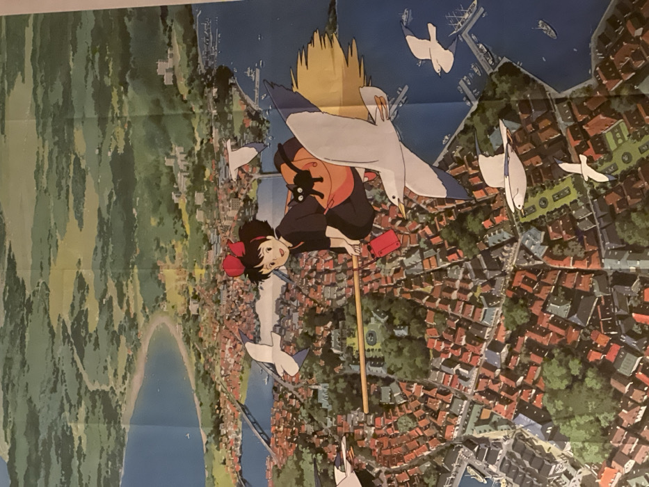 An anime poster depicts a young witch, possibly Kiki, riding a broomstick over a European-style cityscape with red rooftops. The witch wears a dark dress, orange jacket, and a large red bow, accompanied by a small black cat. Several seagulls fly around her. The backdrop includes a harbor with boats, a bridge, and lush green hills fading into the distance. The artwork features soft colors and detailed architecture.