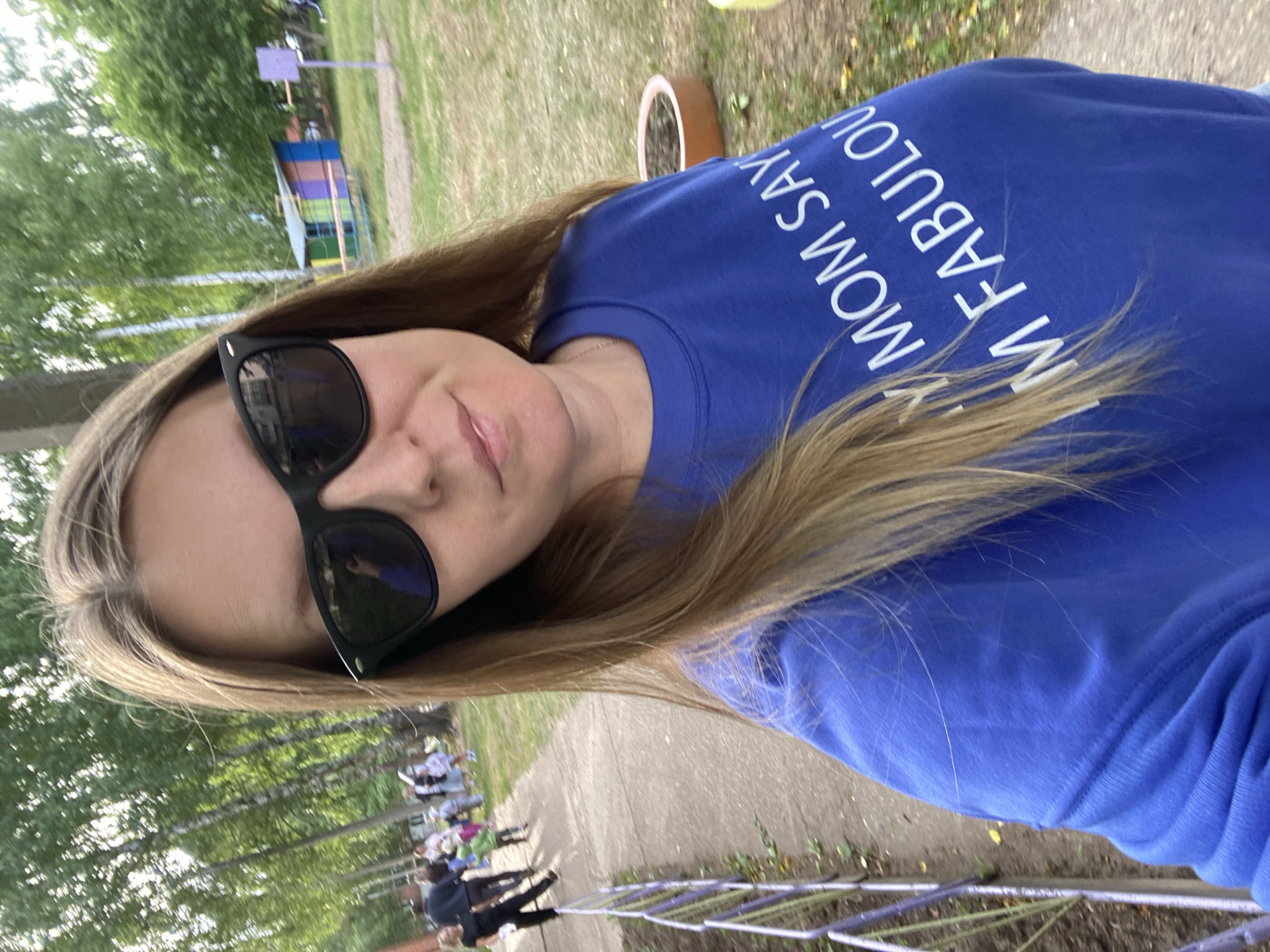 The image shows a woman wearing sunglasses and a blue sweatshirt with the phrase "My Mom Says I'm Fabulous" on it. She is outdoors, likely in a park or recreational area. There is a group of people in the background, seemingly watching a performance or event. The location is specified as the city of Noviy, Russia. Details such as the exact event in the background or specific features that indicate the location within Noviy are not discernible from the provided image.