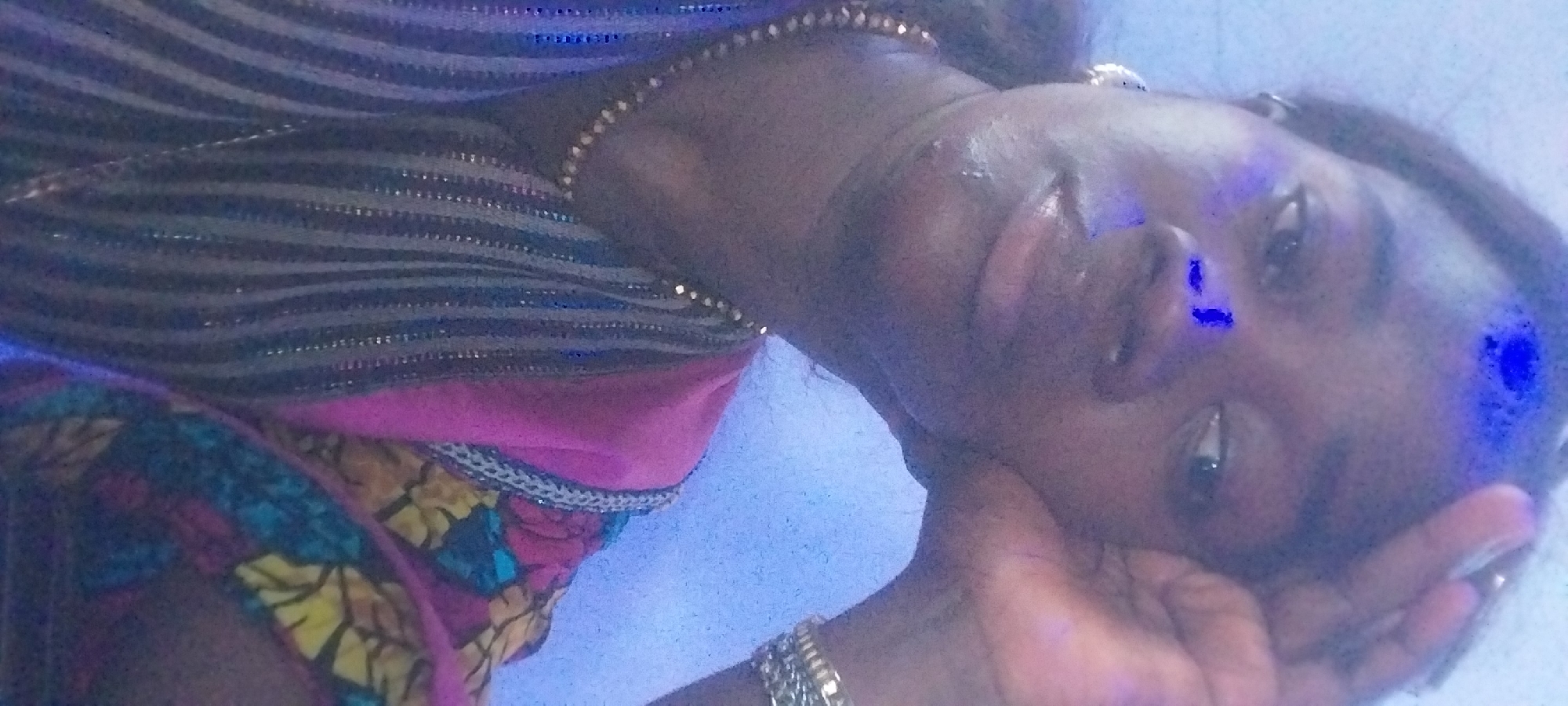 The image is a close-up, self-portrait style photograph of a young Black woman. She is wearing a brightly colored, patterned garment, possibly traditional African attire, with shades of pink, yellow, blue, and black. She also has on a delicate gold necklace. Her right hand is resting on her forehead, and her left arm is bent, with her hand near her face. There are blue light reflections on her forehead and nose, suggesting the presence of a light source with a blue hue. The background is a textured, light blue wall. The woman's expression is somewhat pensive, with her eyes looking slightly off-camera. The image appears to have been taken indoors. The photograph does not provide any specific visual cues to determine its exact location within Akpaka Forest Reserve, Nigeria, as it focuses solely on the subject and her immediate surroundings.