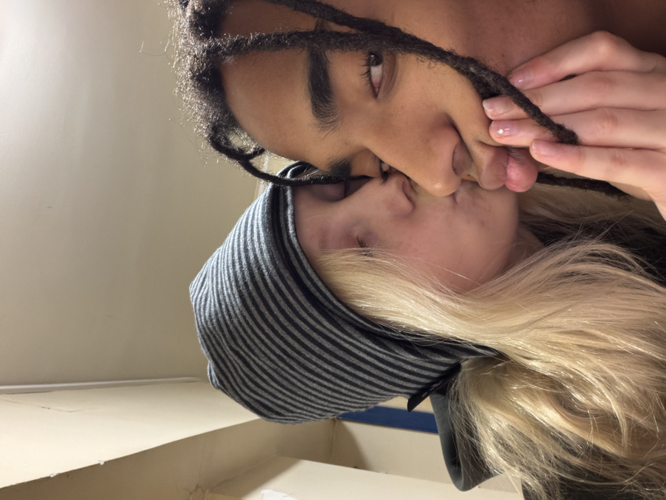 A close-up captures two individuals in an intimate moment, heads tilted. One has dark skin, dreadlocks, and dark brows, with an eye visible. The other has fair skin, blonde hair, and wears a gray-and-black striped beanie. Their lips are pressed together in a kiss. A hand with polished nails is visible near the dreadlocks. The background shows a plain wall.