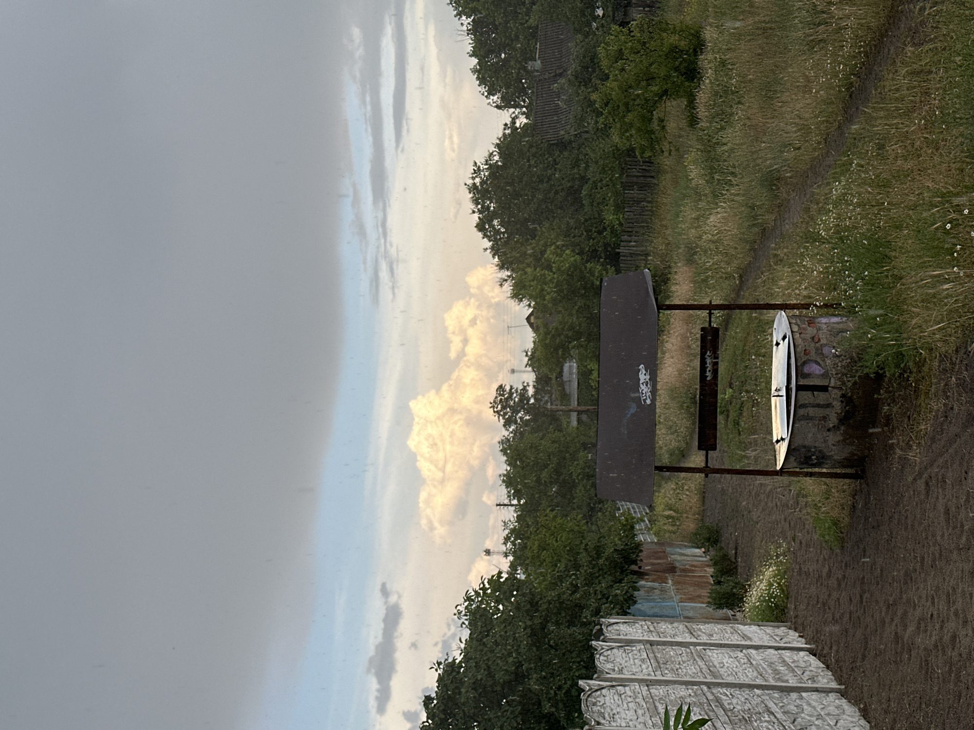 A well with a metal roof stands in the foreground of this outdoor scene, in what appears to be a rural or semi-rural setting. The well is constructed of concrete rings with some graffiti on its surface. A metal crank mechanism is attached to the roof structure. A dirt path leads away from the well towards the background. To the left of the path is a concrete fence, and beyond that, some corrugated metal fencing. Lush green trees and vegetation are abundant, with a field of tall grass and small white flowers bordering the path to the right. In the background, the sky is filled with dramatic, heavy clouds, with a bright, illuminated cloud formation in the center. It appears to be raining, as evidenced by streaks visible in the image. The overall atmosphere is overcast and somewhat moody.