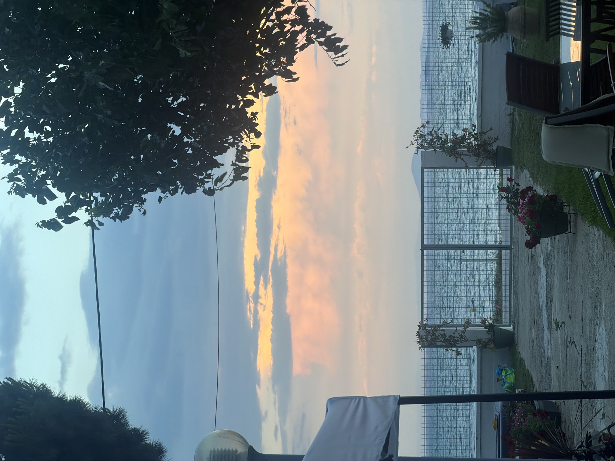 The image depicts an outdoor scene with a view of the sea at sunset. In the foreground, there is a paved area with some potted plants and outdoor furniture, including chairs and a table. A white canopy structure is partially visible on the left. A metal fence separates the property from the sea. The sea itself appears calm with gentle waves.
In the background, the sky is filled with dramatic clouds illuminated by the setting sun, casting hues of orange, pink, and yellow. A distant landmass or island is visible on the horizon. The overall atmosphere suggests a tranquil evening by the coast.
The image does not provide specific details about the city of Mandra, Greece, beyond its geographical context as a coastal location. There are no people present in the image. The scene is static and serene.