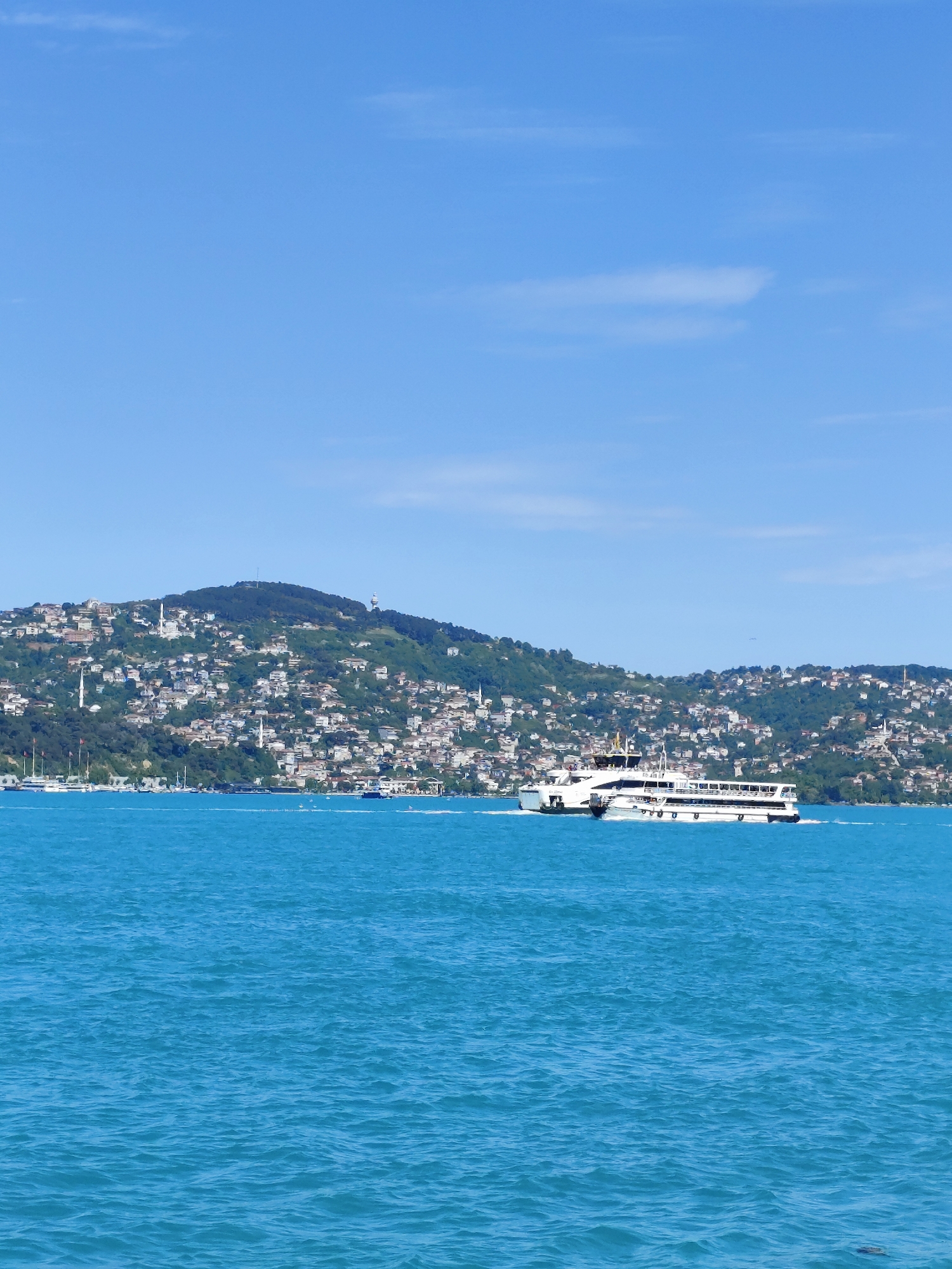 The image depicts a sunny, daytime scene on the Bosphorus strait in Sarıyer, Turkey. A white ferry with multiple decks traverses the blue water, creating a wake behind it. The coastline features a densely populated hillside covered with buildings and greenery, with a radio tower on the highest point of the hill. The sky is mostly clear blue with some sparse white clouds.