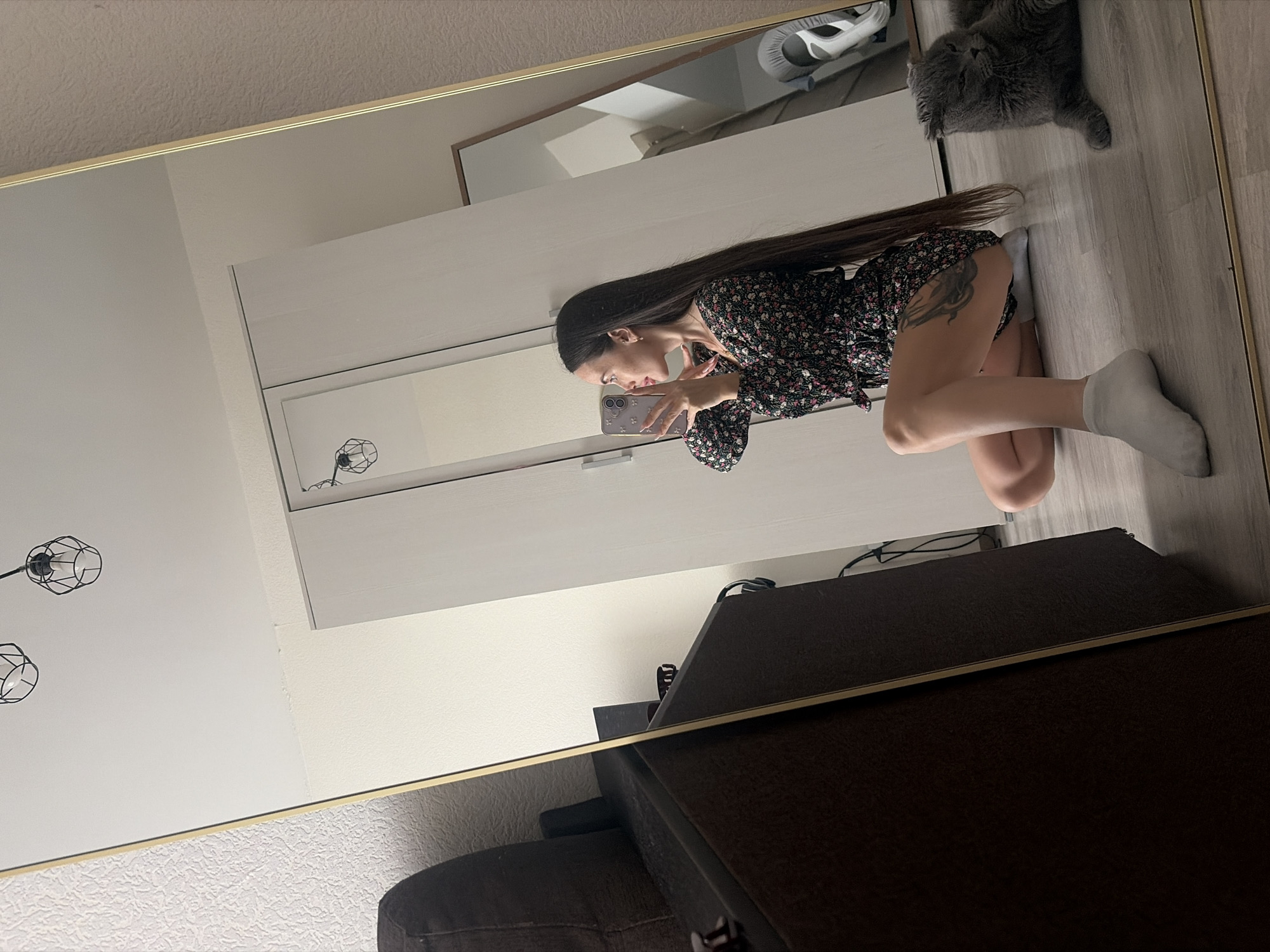 This indoor mirror selfie, rotated 90 degrees counter-clockwise, captures a young woman kneeling on a light wooden floor. She has long dark hair and is wearing a dark floral mini-dress with white socks. She holds a light-colored smartphone, possibly an iPhone, to take the picture, appearing focused on its screen. A tattoo is visible on her upper right thigh.
In the reflected background, a fluffy grey cat rests on the floor, looking towards the camera, alongside a pair of athletic sneakers. Behind the woman, white wardrobes or cupboards with horizontal handles are visible in the reflection. A dark upholstered sofa or armchair is partially seen to the right. The walls are light-colored, and the ceiling (at the bottom of the original image) features textured wallpaper. Two black, cage-style pendant lights are also reflected on the left side of the mirror.
The scene depicts a modern home interior, consistent with a residential setting in Saint Petersburg, Russia. The time of day is ambiguous due to indoor lighting. There is no visible text in the image.
