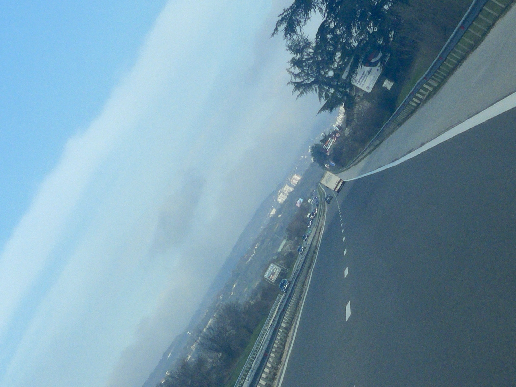 The image depicts a view from a vehicle traveling on a highway near Ignatievo, Bulgaria. The highway curves slightly to the right. A line of vehicles is visible in the distance on the opposite side of the road. A crash barrier runs along the right side of the road. A tree is partially visible in the upper right corner, near a road sign. A town and landscape are visible in the background under a cloudy sky.