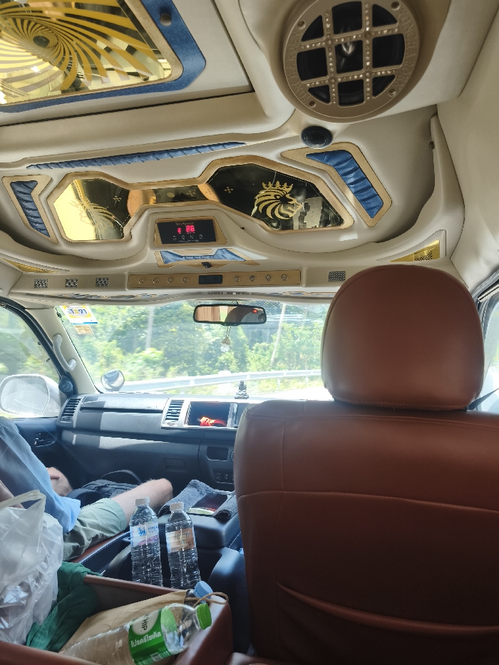 The image captures the interior of a customized passenger vehicle, likely a van or bus, in transit through Ban Wang Pra Chan, Thailand. The viewpoint is from a rear seat, looking forward towards the front cabin. The vehicle's ceiling is elaborately decorated with cream-colored upholstery, gold-colored trim, and blue textured accents. Overhead features include gold-mirrored panels, one displaying a crowned lion emblem, a large spiral design in gold, and a central digital display showing "88" in red, along with a clock icon. A circular speaker grille is visible in the upper right.
In the immediate foreground, a brown leather headrest occupies the right side of the frame. Forward of this, the lower torso and legs of a passenger, wearing light-colored shorts, are visible in the front passenger seat. Between the front seats, on a central console, two clear plastic bottles of Singha R-O drinking water stand upright, next to a dark smartphone or tablet resting on a grey cloth. A dark bag is positioned on the floor or console area in front of the passenger. In the lower left, a white plastic bag and a green-capped "TIP" brand water bottle are contained within a brown receptacle.
Through the front windshield, a multi-lane road with a white metal guardrail is visible, bordered by dense green foliage and trees under a bright daytime sky. A small, dark figurine, possibly a Buddha statue, is present on the dashboard. A rearview mirror hangs centrally, with a small decorative item suspended from it. The dashboard appears standard for a commercial van.